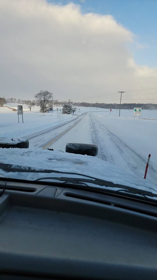 Snowy rural road seen from inside a vehicle, with tire tracks and snow-covered fields ahead.