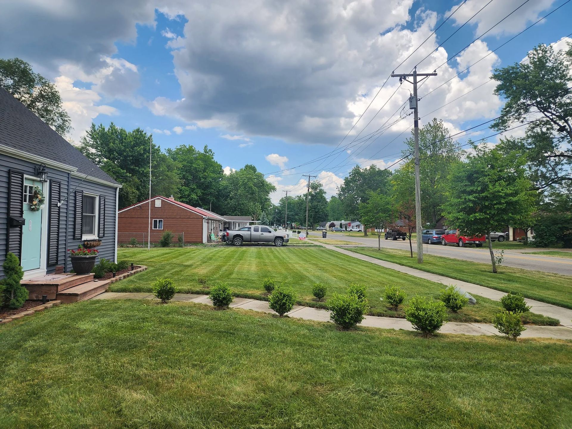 Suburban street with green lawn, small houses, trees, and a partly cloudy sky