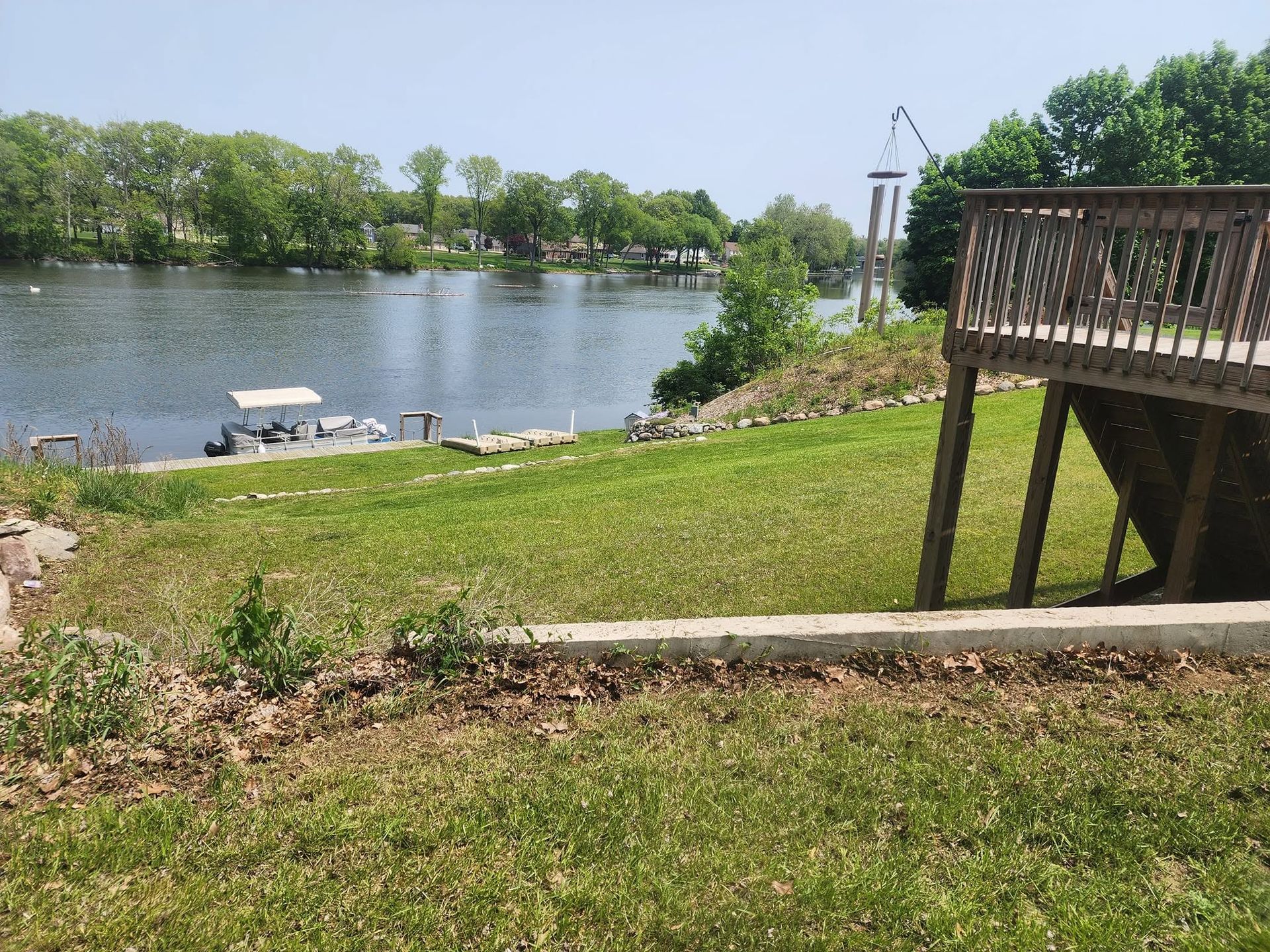 Grassy riverbank with a dock, trees across the water, and a wooden deck railing on the right.