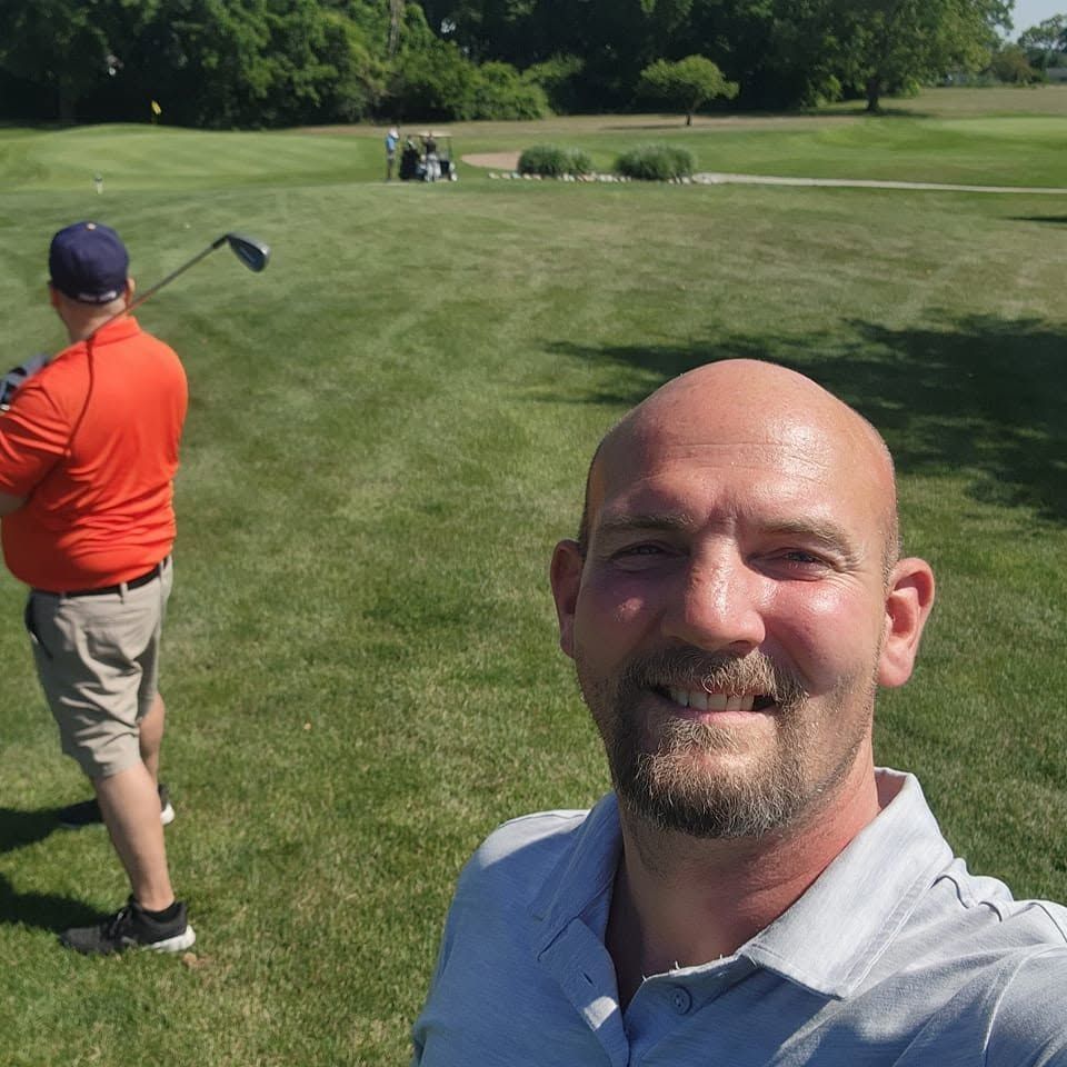 Two men on a golf course, one taking a selfie in the foreground and another swinging a club in the background.