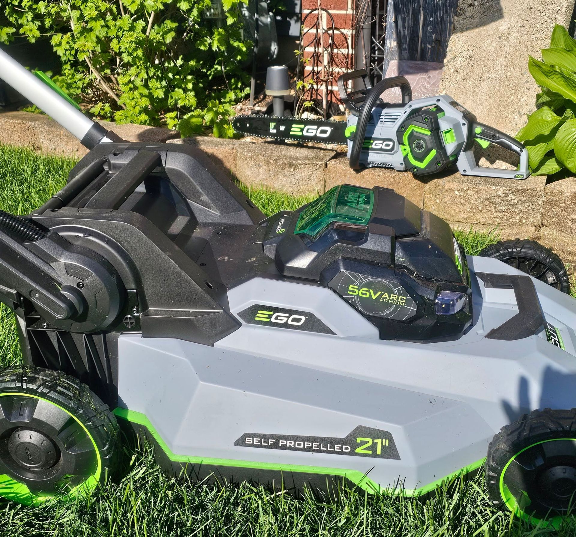 Green EGO lawn mower and trimmer on grass beside a hedge and patio path