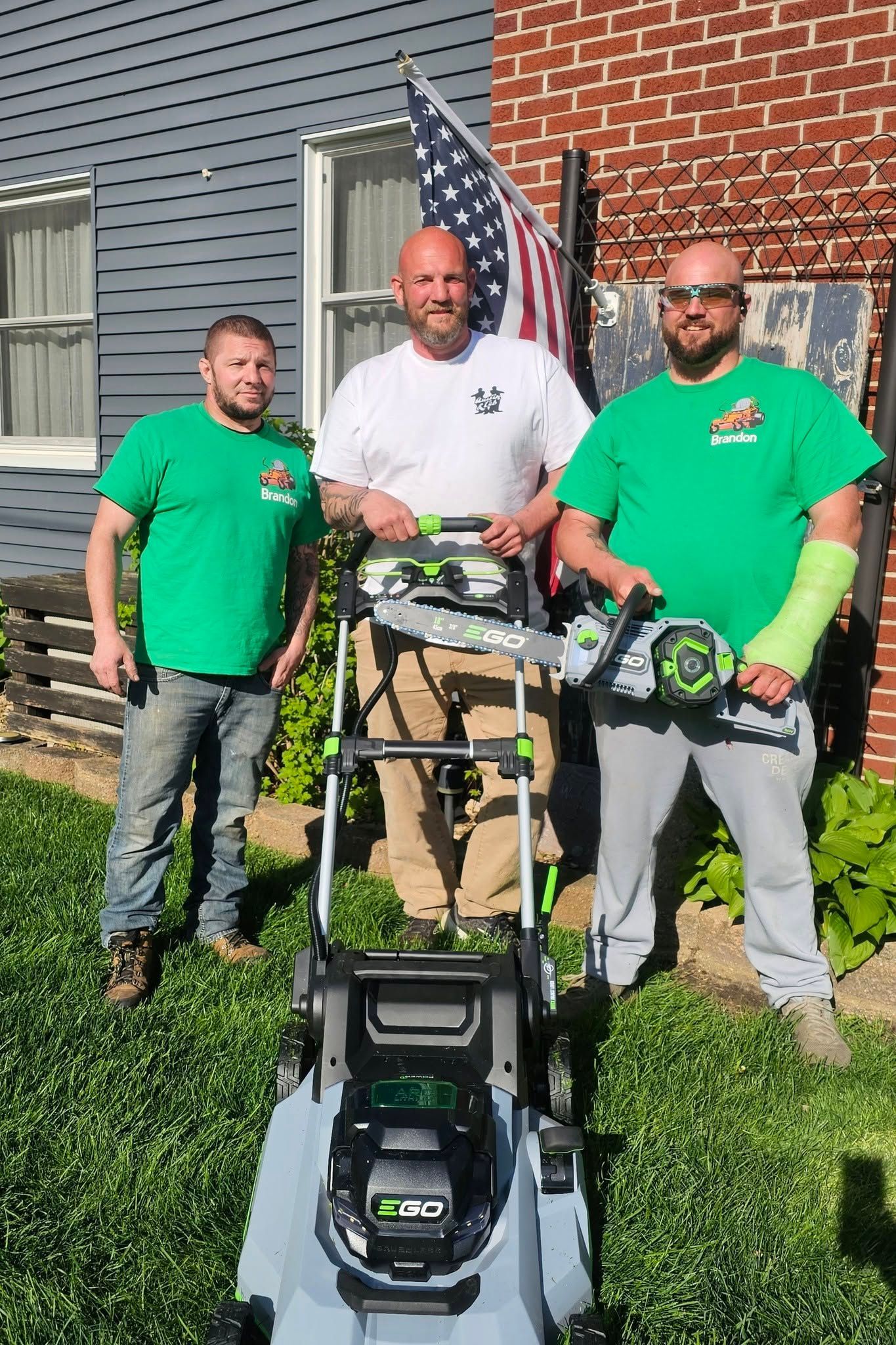 Three workers in green shirts stand behind a lawn mower in front of a brick building and American flag.