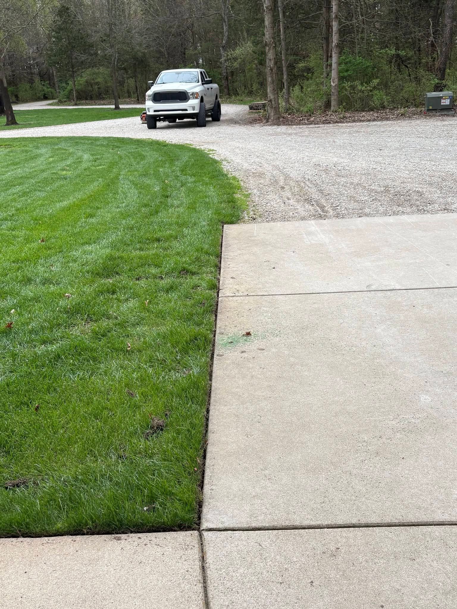 Driveway with concrete pad, green lawn, and a white truck on a gravel road in the background