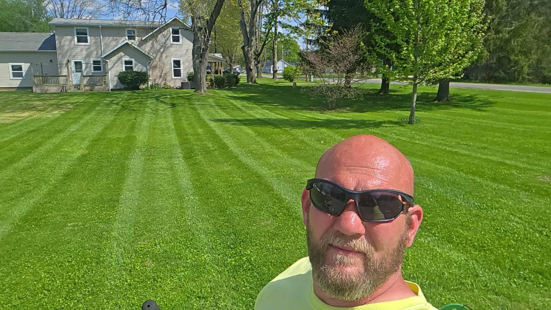 Man in sunglasses taking a selfie in front of a large house and neatly striped green lawn