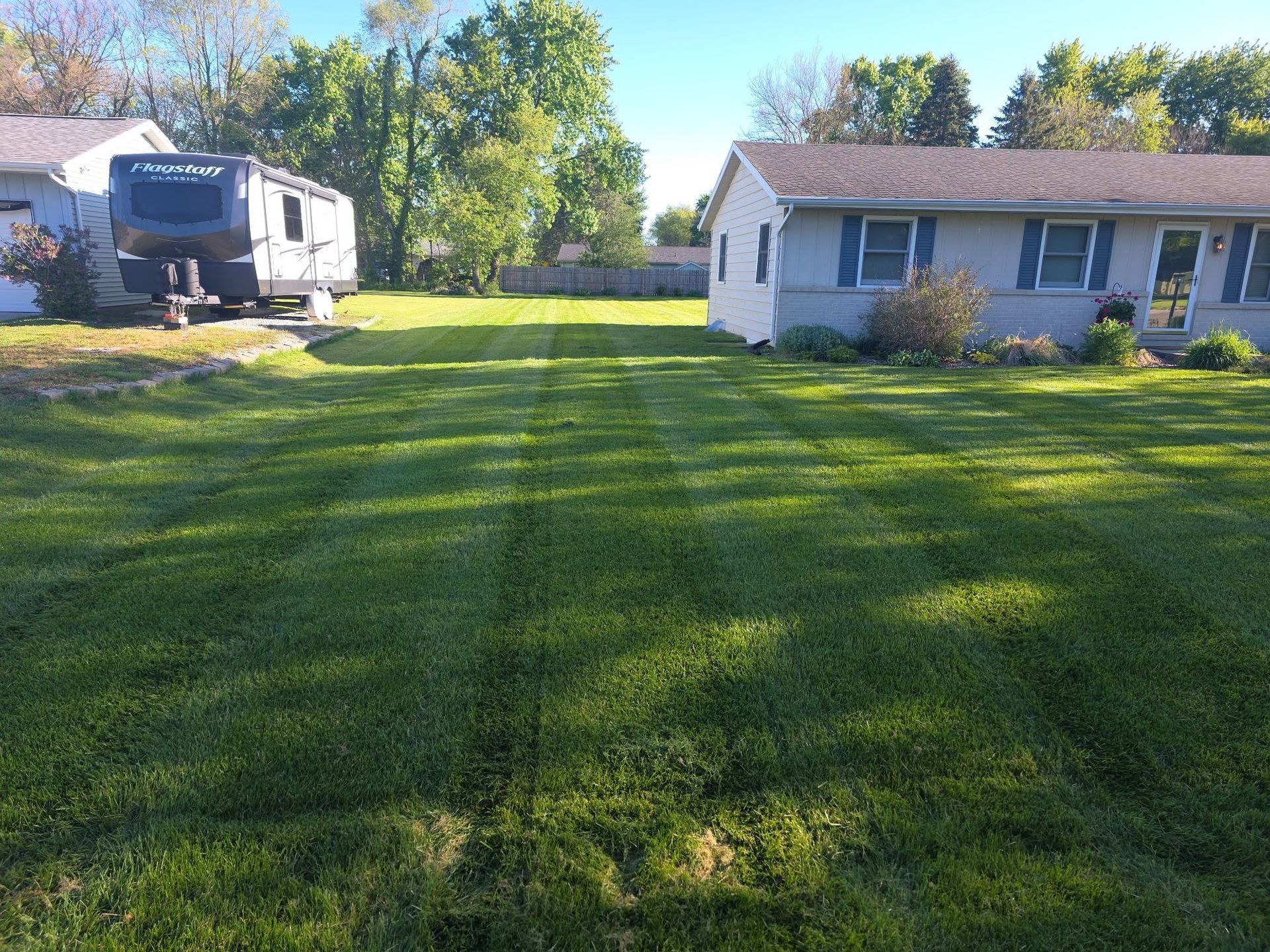 Grassy suburban yard with a white house, RV, and trees in bright sunlight