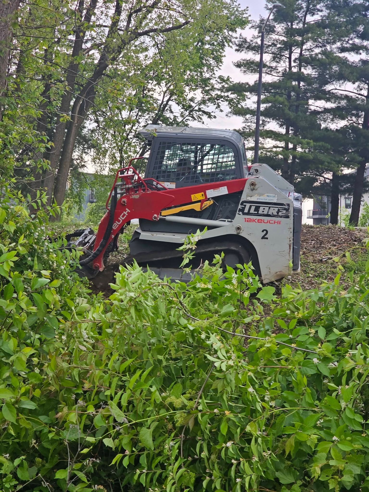Red and gray Bobcat skid-steer on a muddy path, surrounded by dense green brush and trees