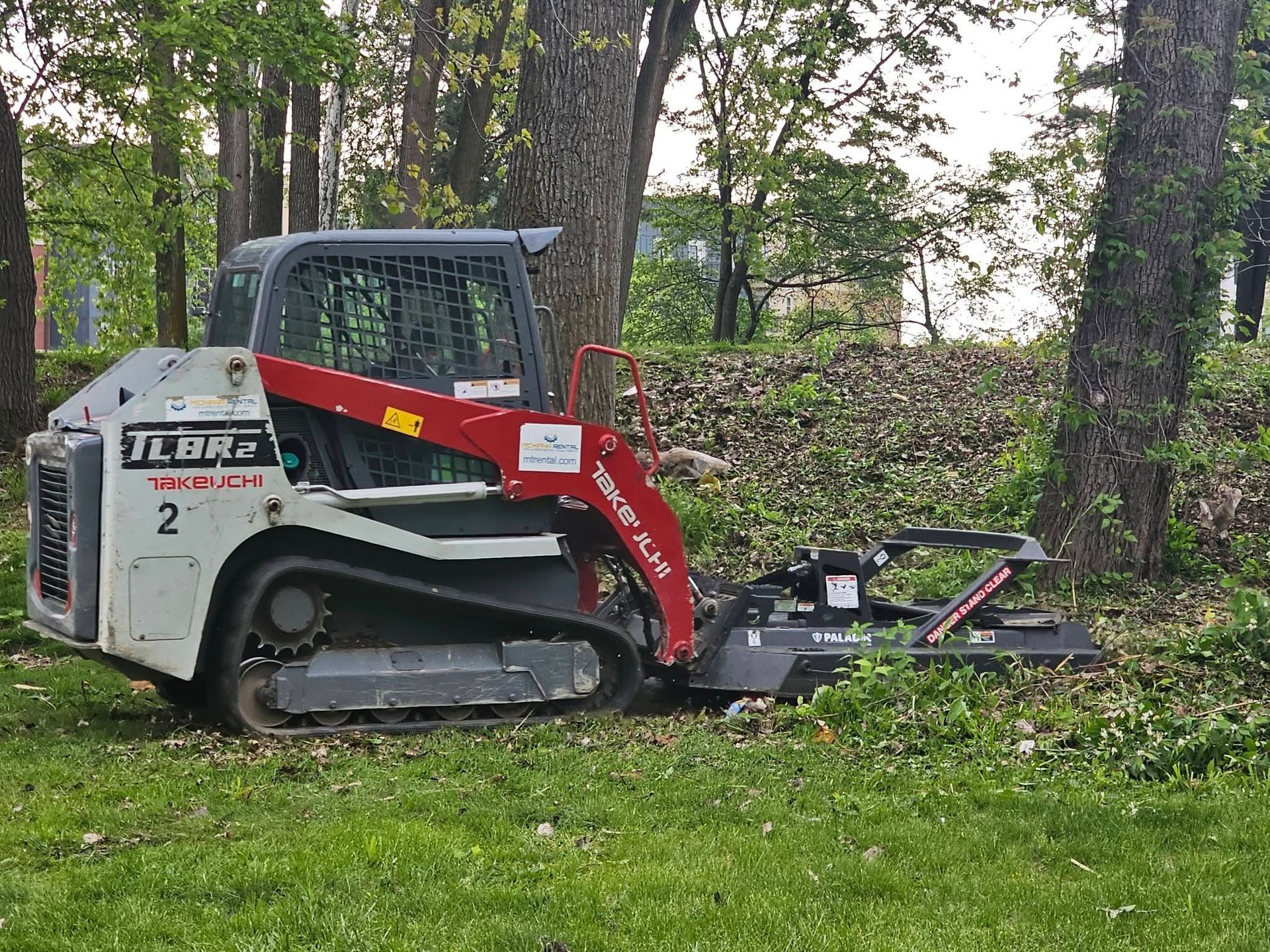 Compact track loader in a wooded area, clearing brush with a red attachment.
