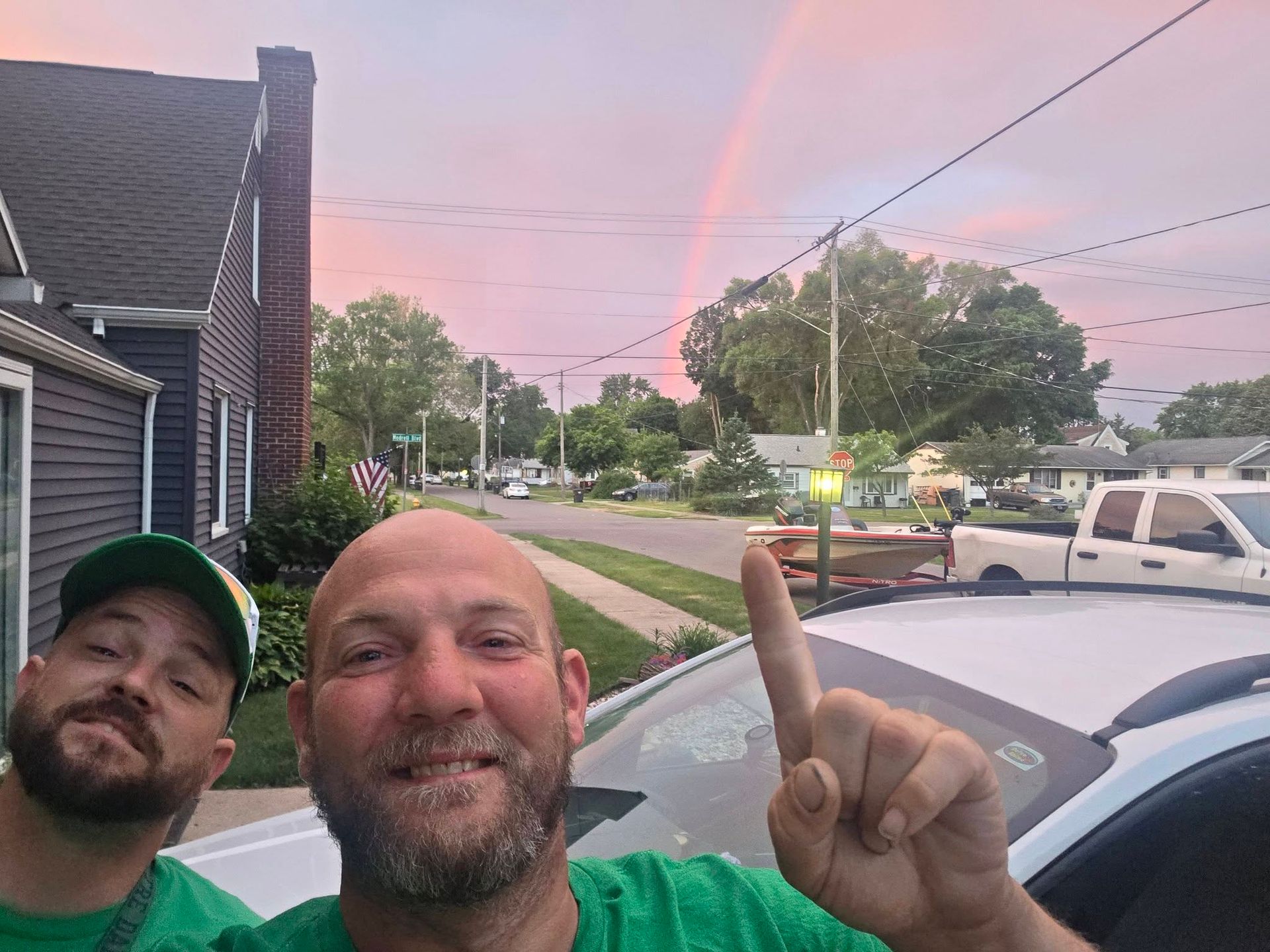 Two men taking a selfie near a white car at sunset, pointing at a pink rainbow in the sky.