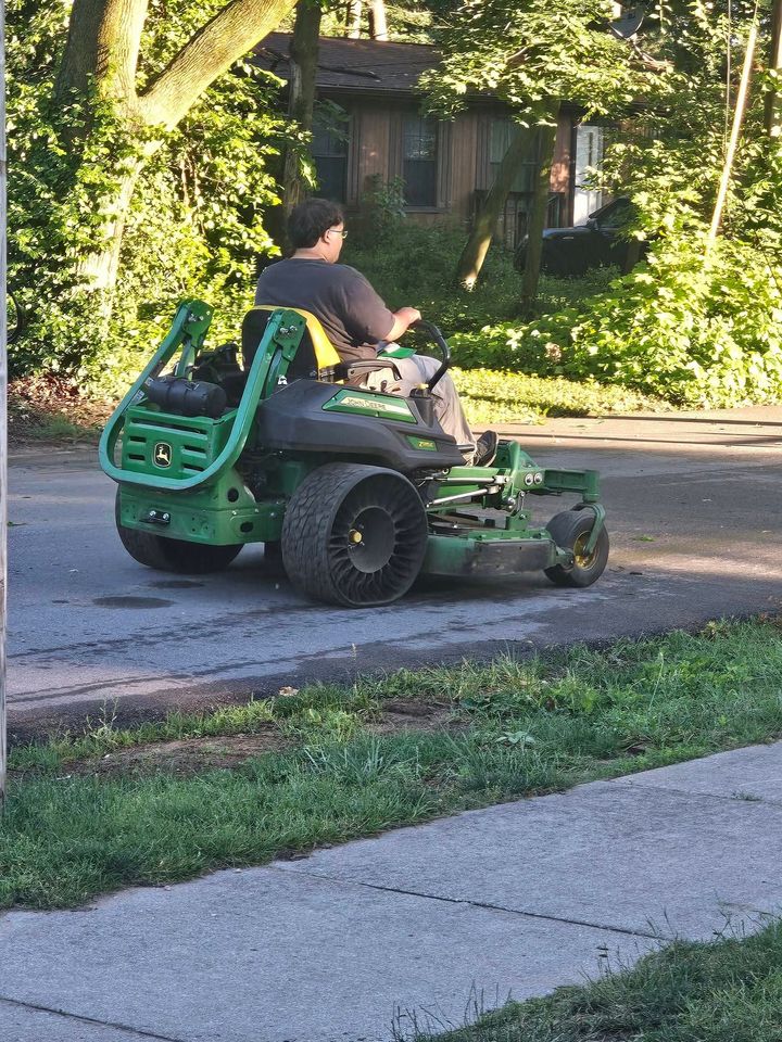 Person riding a green lawn mower on a street beside a sidewalk and trees