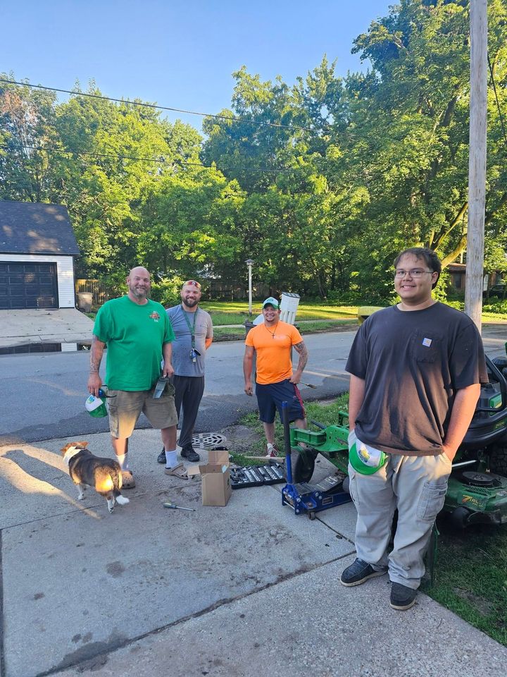 Four people standing outdoors near a grill and bike on a sidewalk by a house, with trees in the background
