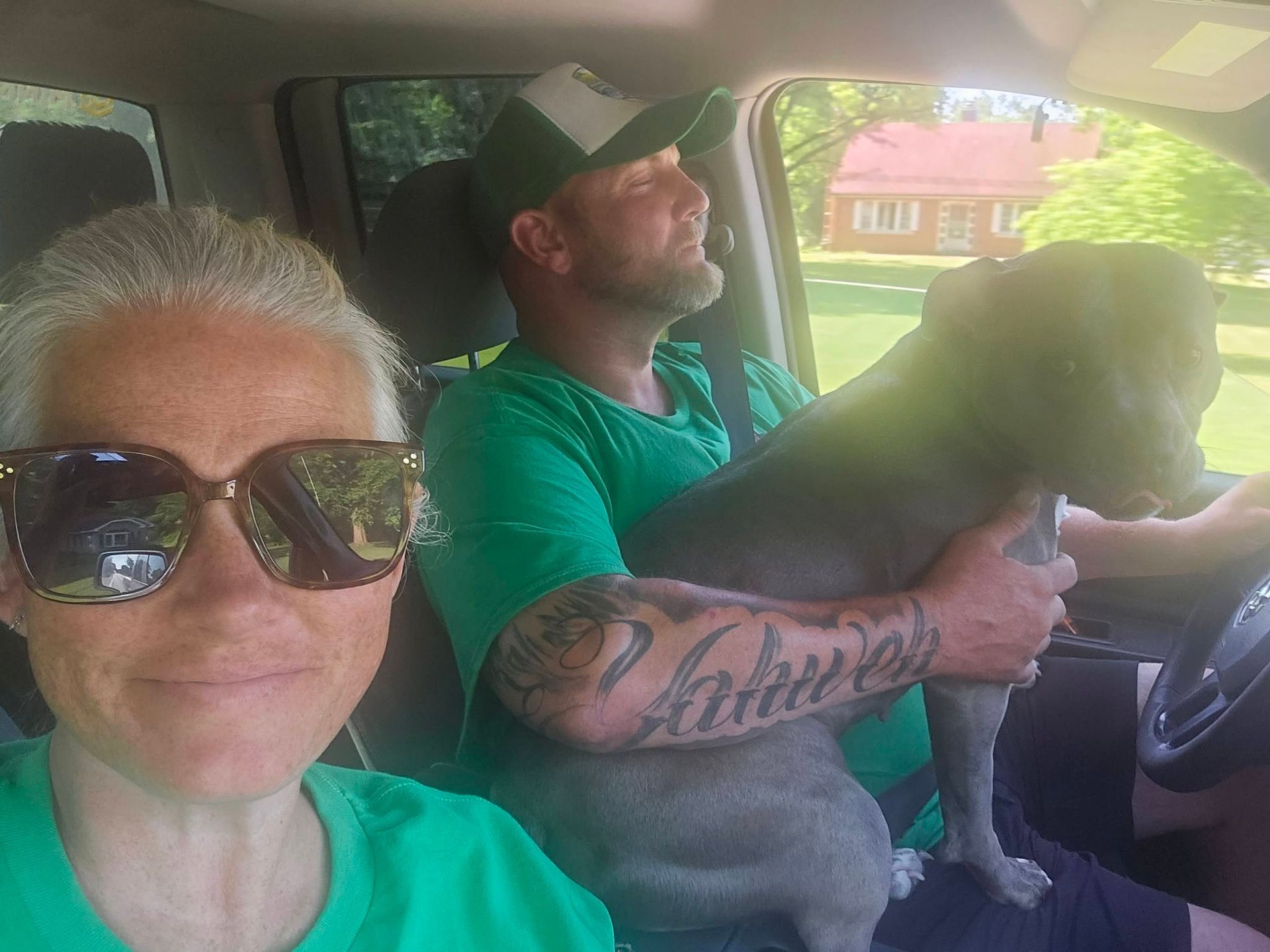 Two people and a dog in a car, one wearing sunglasses and the other driving in a green shirt.