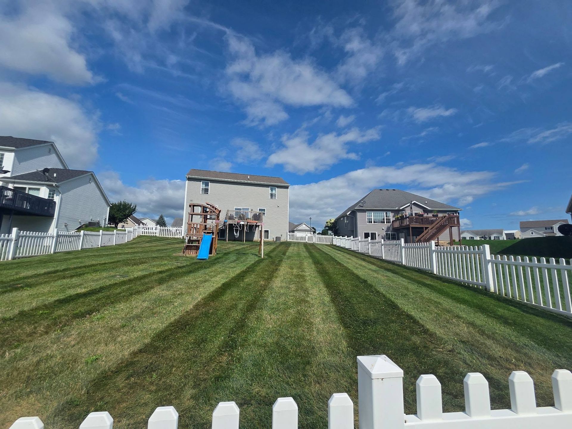 Grassy lawn with white fencing leading to beach houses under a blue, partly cloudy sky