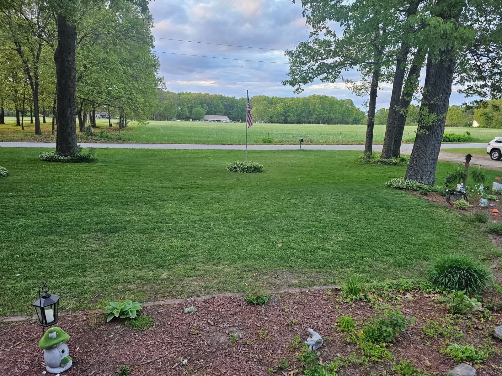 Grassy front yard with trees, flower bed, and a road in the background under a cloudy sky