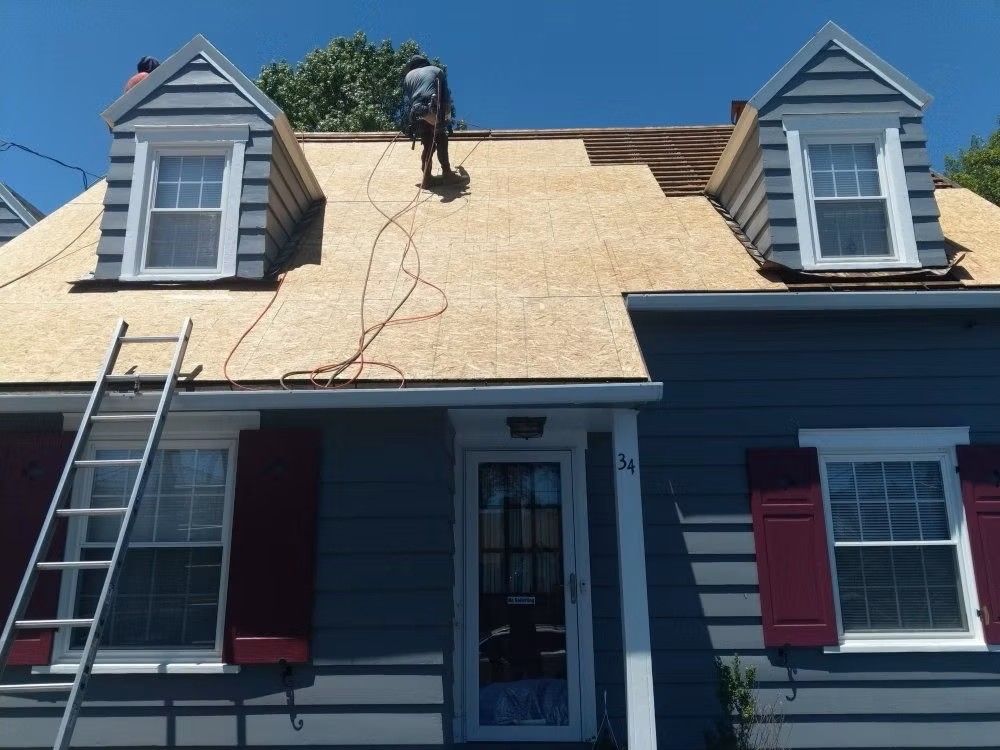 Worker repairing roof on blue house with two dormer windows and a ladder against the front.