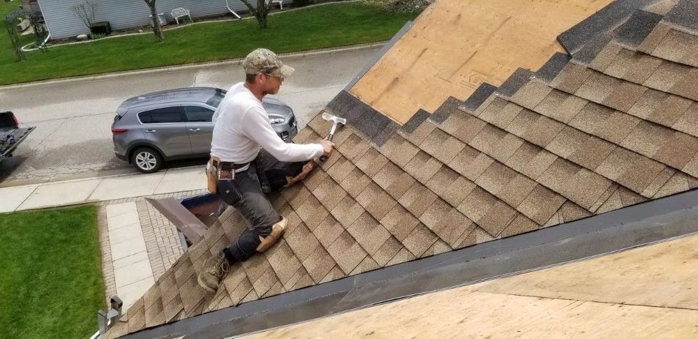 Worker on a steep shingled roof inspecting or repairing the surface near a residential street.
