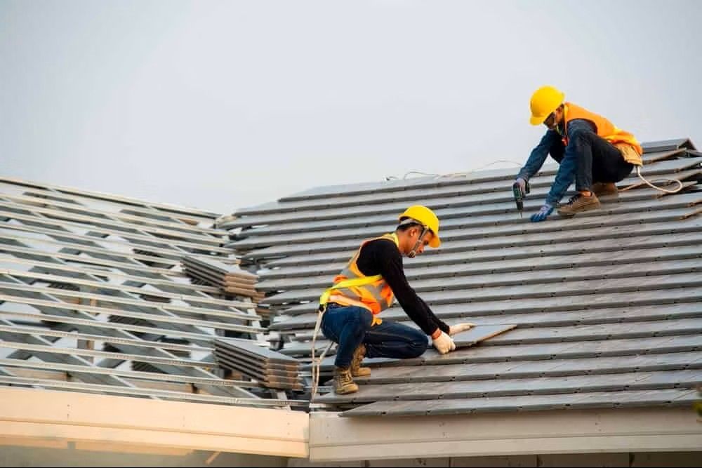 Two workers in hard hats repairing a gray roof with exposed damaged sections