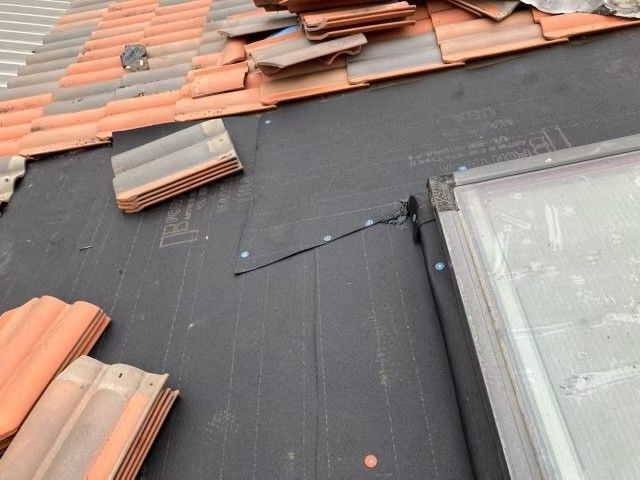 Roof under repair with black underlayment, stacked terracotta tiles, and a skylight window