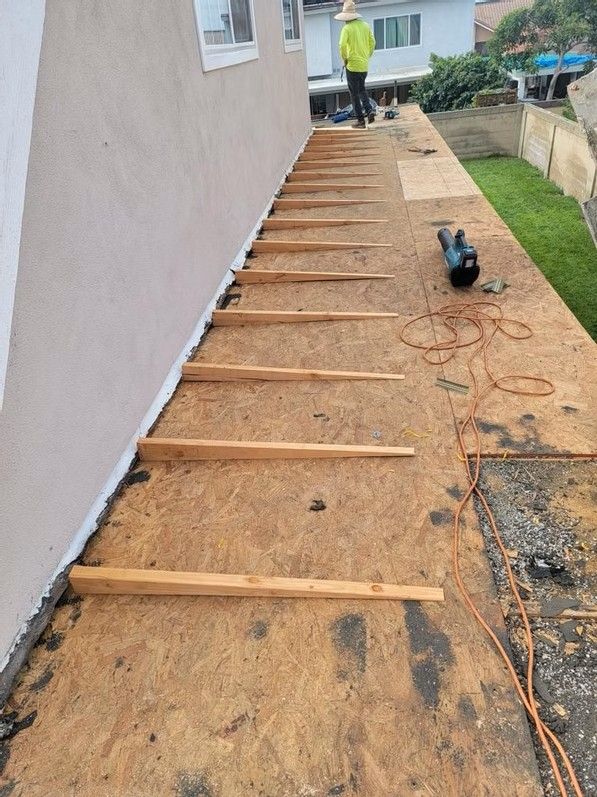 Outdoor construction site beside a house with wooden joists laid over gravel and tools scattered along the path