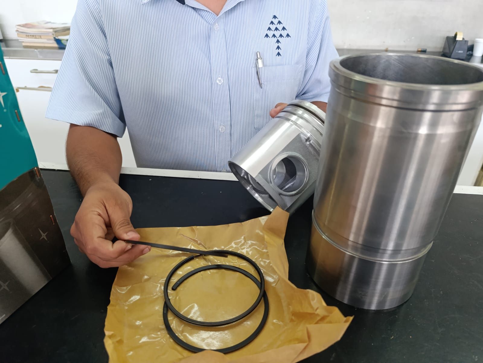 Persona sosteniendo anillos de pistón, con un pistón y una camisa de cilindro sobre una mesa.