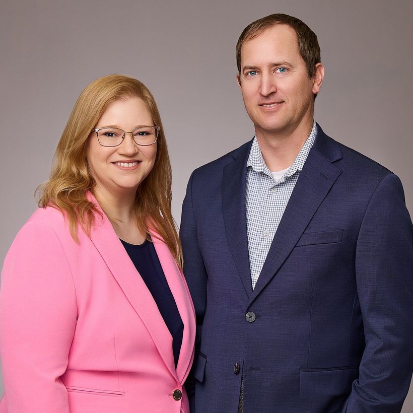 Two people posing against a plain gray backdrop, wearing pink and navy blazers, smiling at the camera