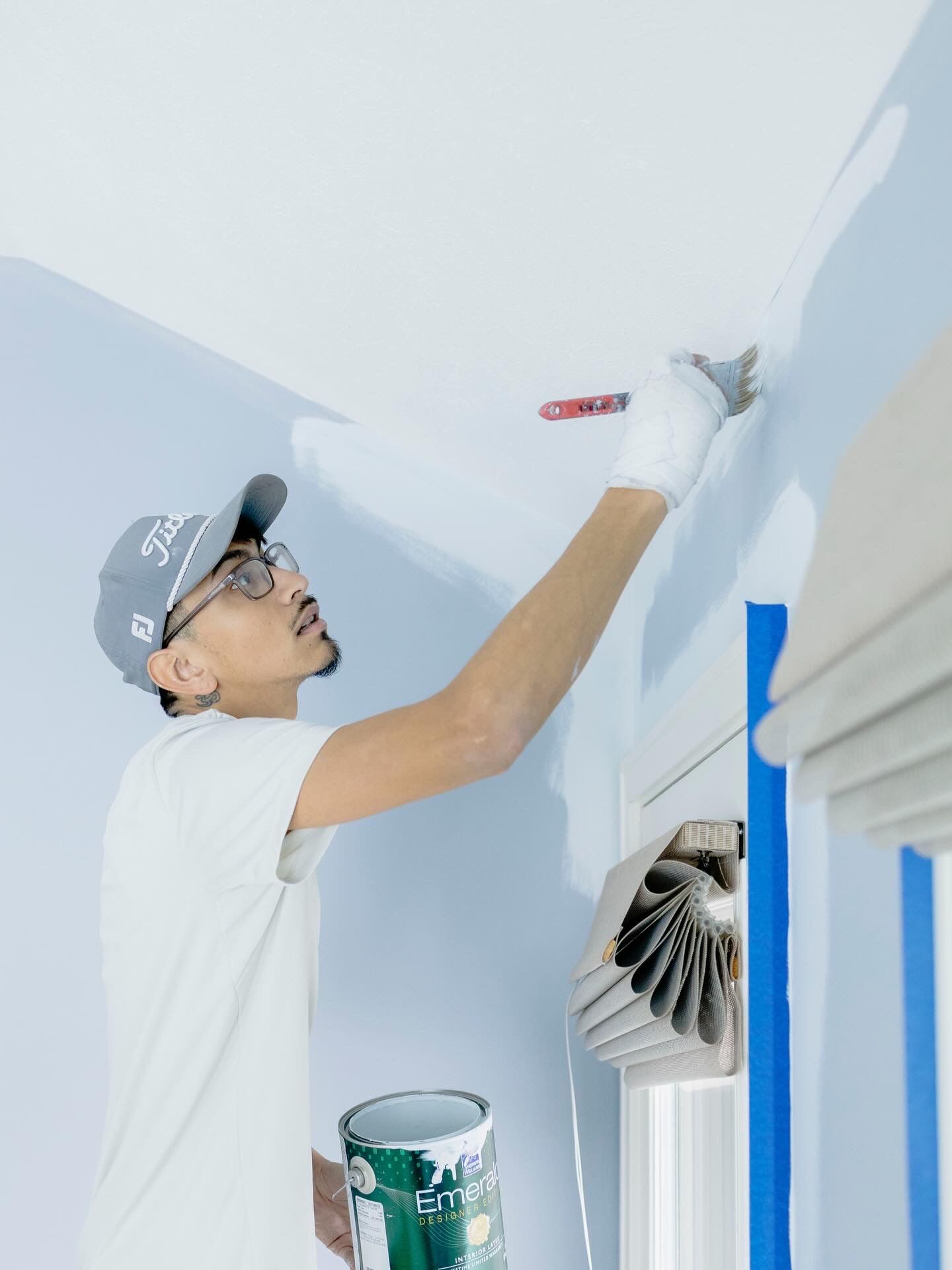 A person wearing a cap and glasses paints the upper edge of a wall white with a brush while holding a paint can.