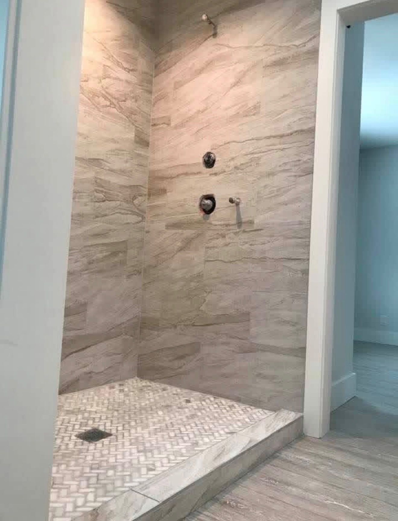 A walk-in shower with light tan stone-look wall tiles, a woven-patterned tile floor, and chrome fixtures.