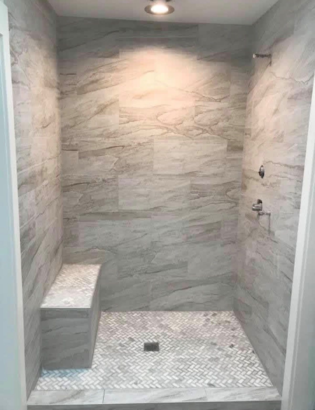 A walk-in shower with light grey stone walls, a built-in bench, and a textured white mosaic tile floor.
