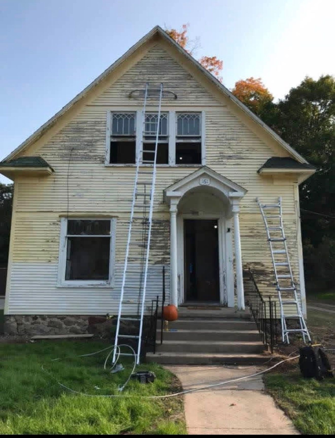 A weathered yellow two-story house with two ladders against the facade, indicating ongoing exterior renovation work.