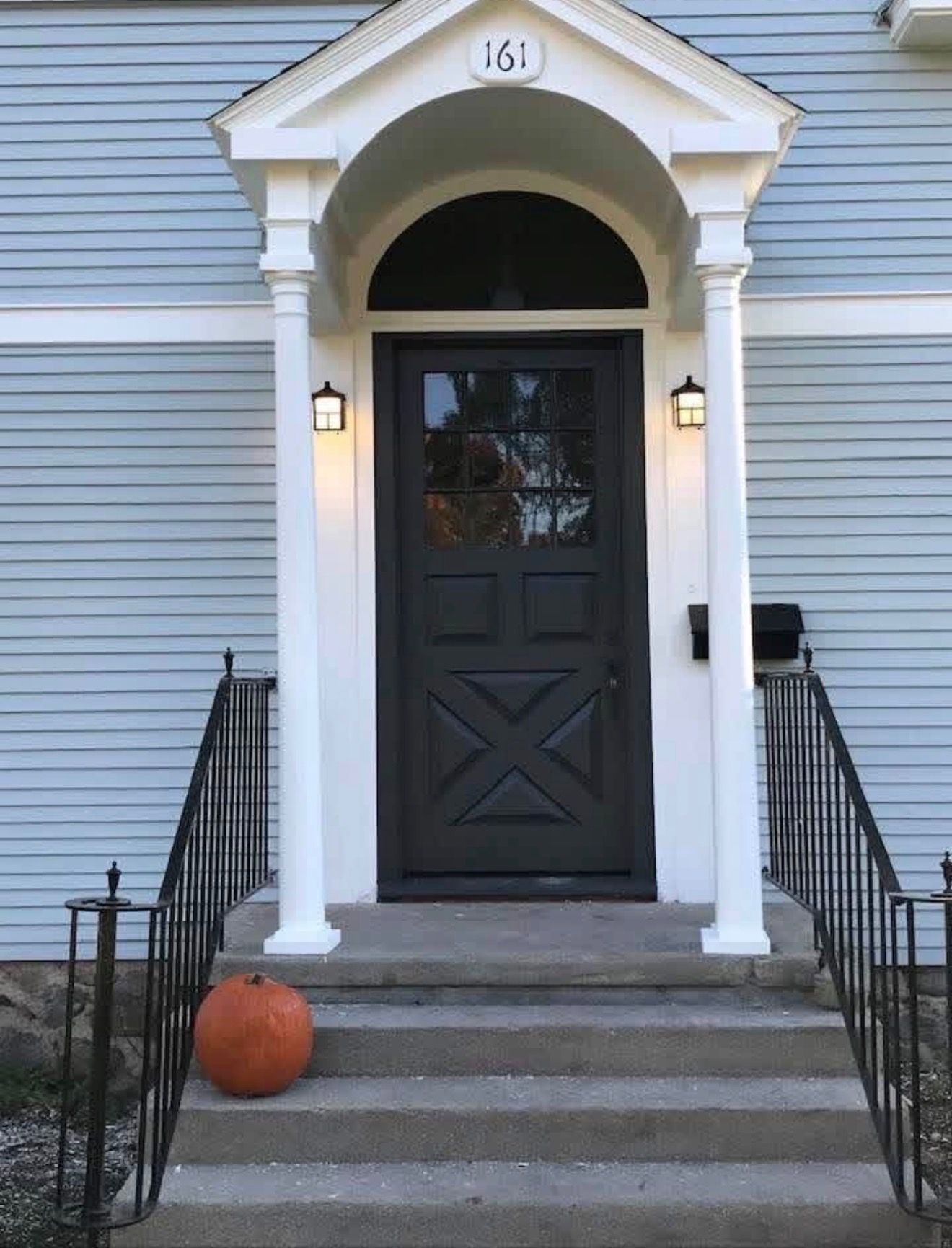 A light blue house front featuring a black door, a white archway, metal railings, and an orange pumpkin on the steps.