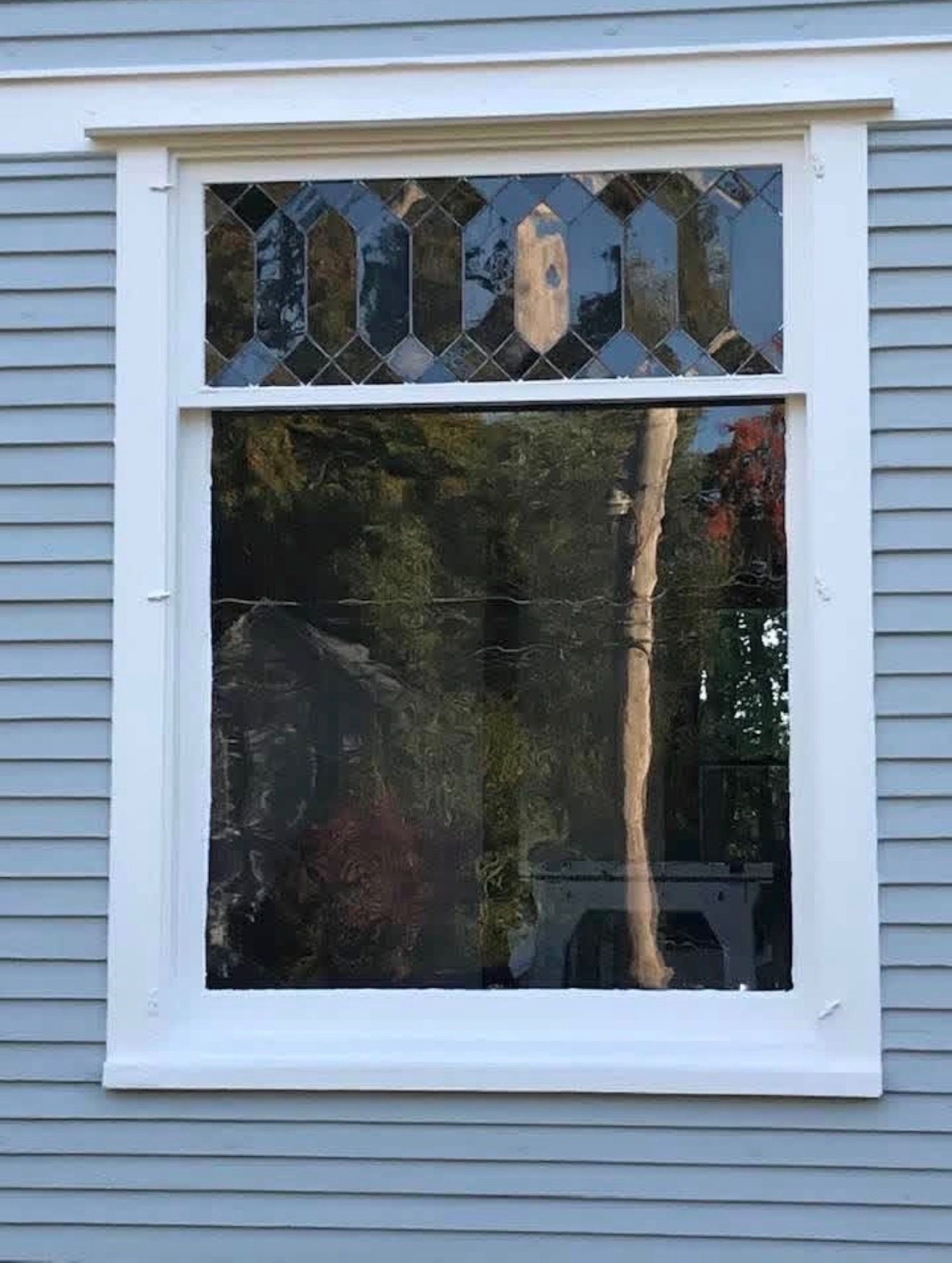 A rectangular window with a geometric stained-glass transom above a large clear pane, set in light blue siding.