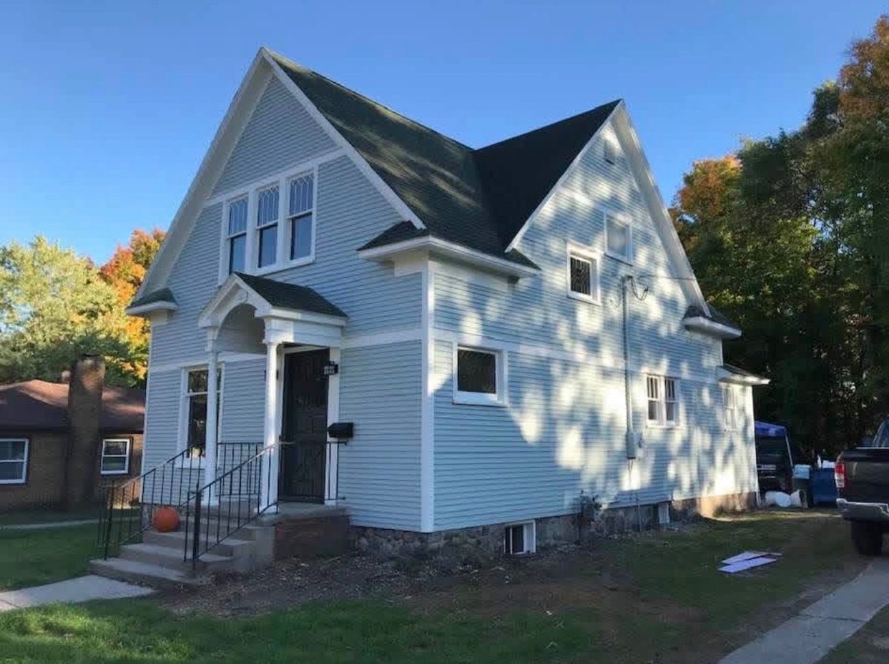 A two-story light blue house with a steep gable roof, white trim, and a covered front porch, set on a grassy lot.