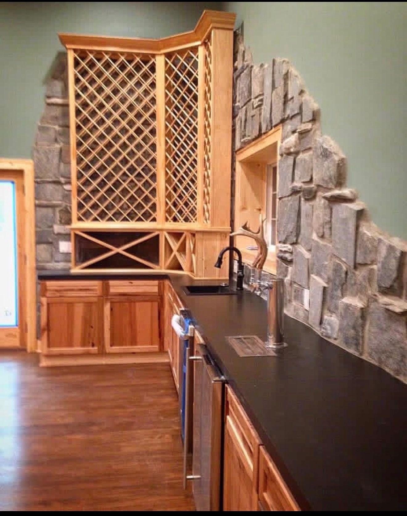 A home bar with wood cabinets, black countertops, a wine rack, and a natural stone backsplash on a green wall.