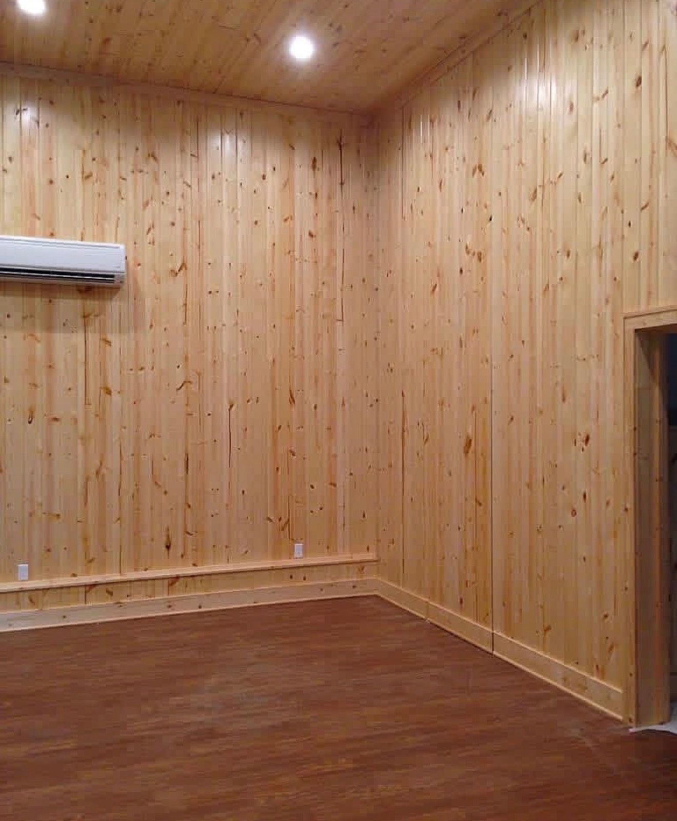An empty room with vertical knotty pine wall paneling, a wood-look floor, recessed ceiling lights, and an AC unit.
