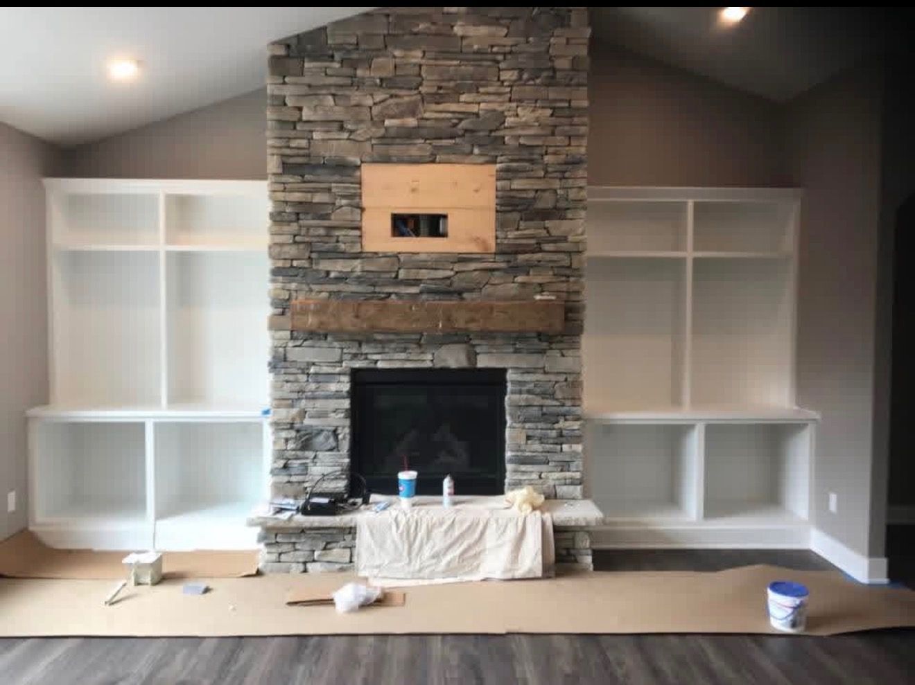 A fireplace with a stone chimney and wooden mantel, flanked by white built-in shelving units in a room under construction.