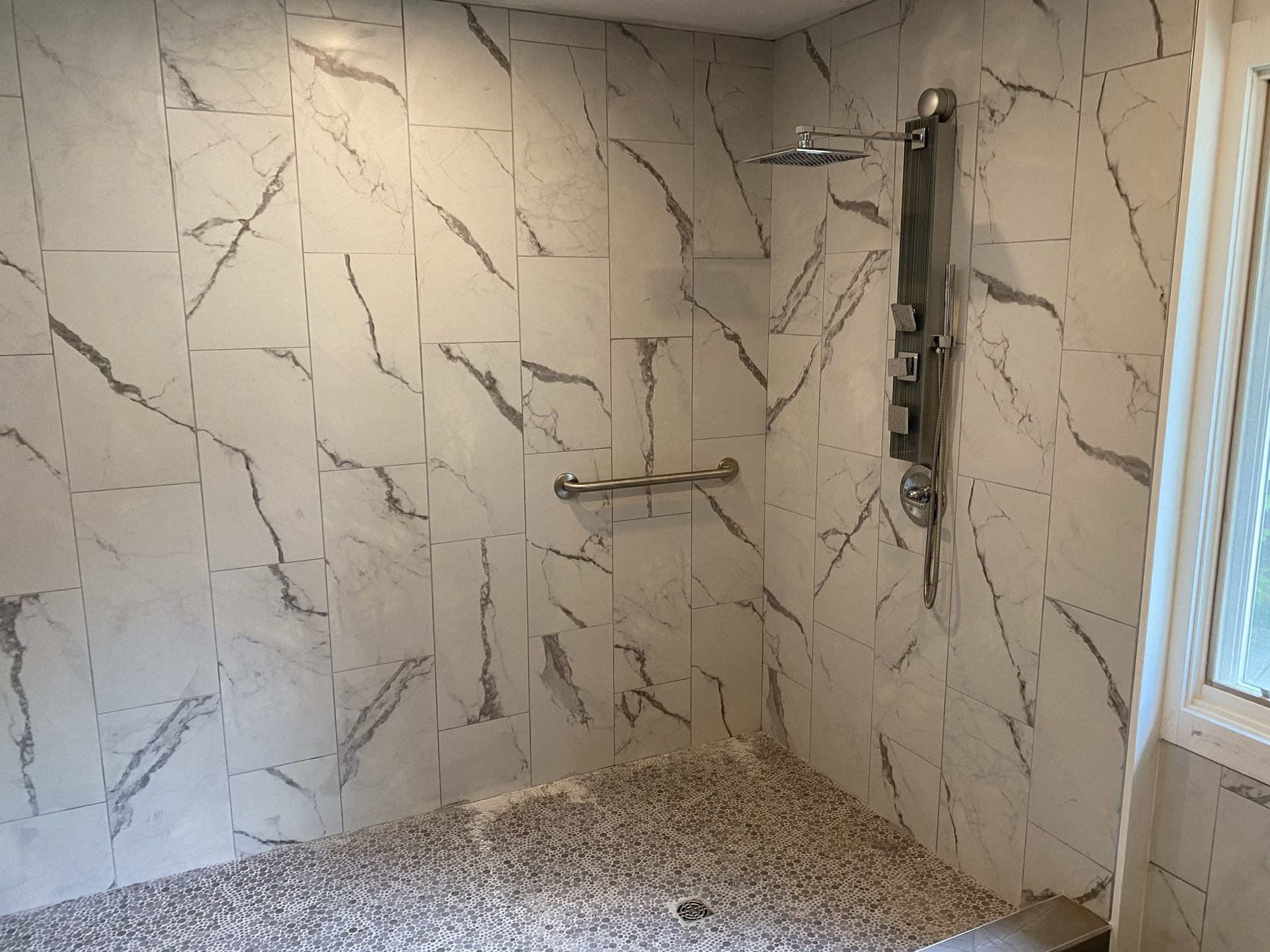 A walk-in shower with white marbled tile walls, a pebble floor, a vertical shower panel, and a mounted grab bar.