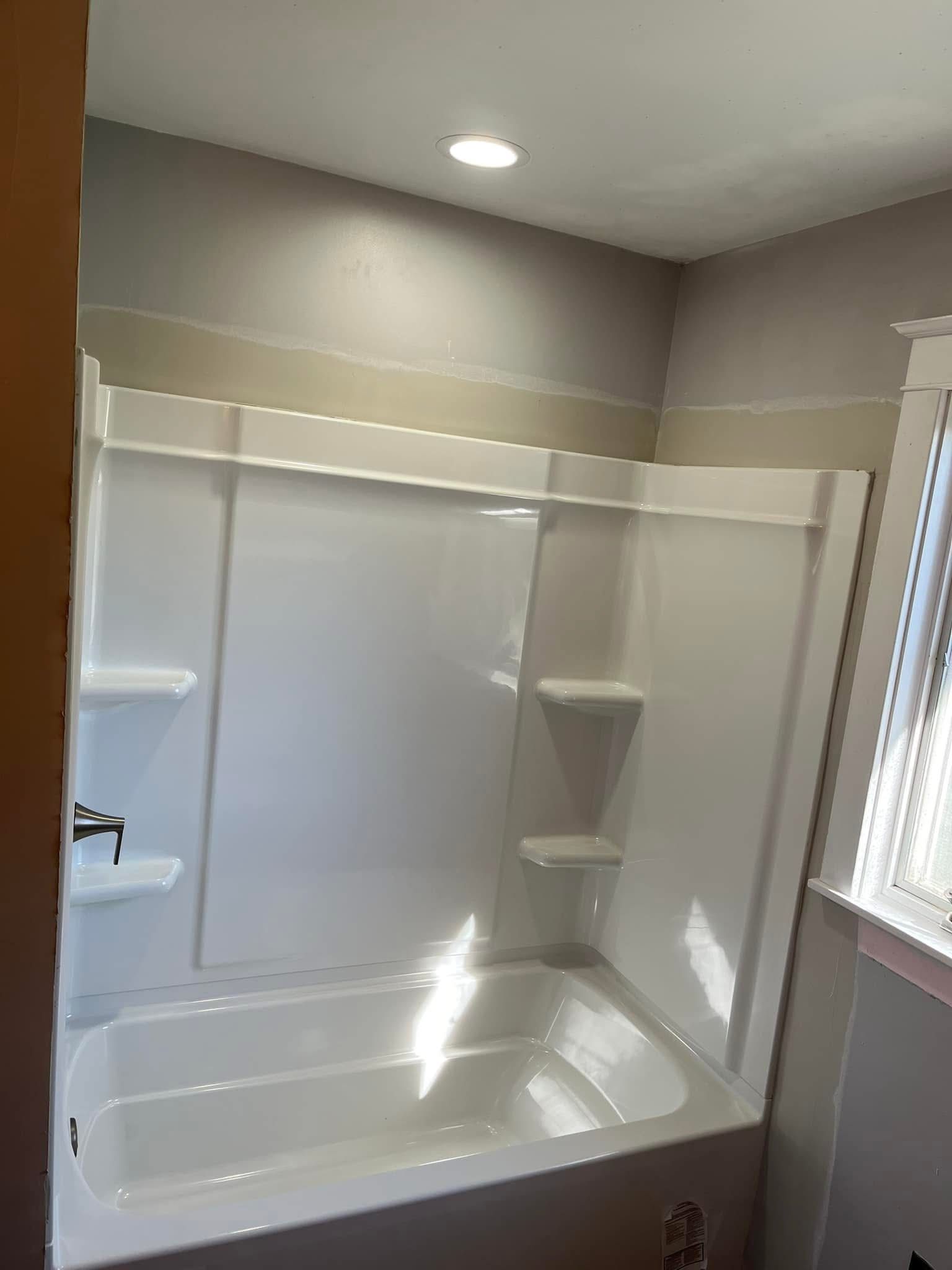 A white bathtub and shower surround with built-in shelves installed against a beige wall in a bathroom.