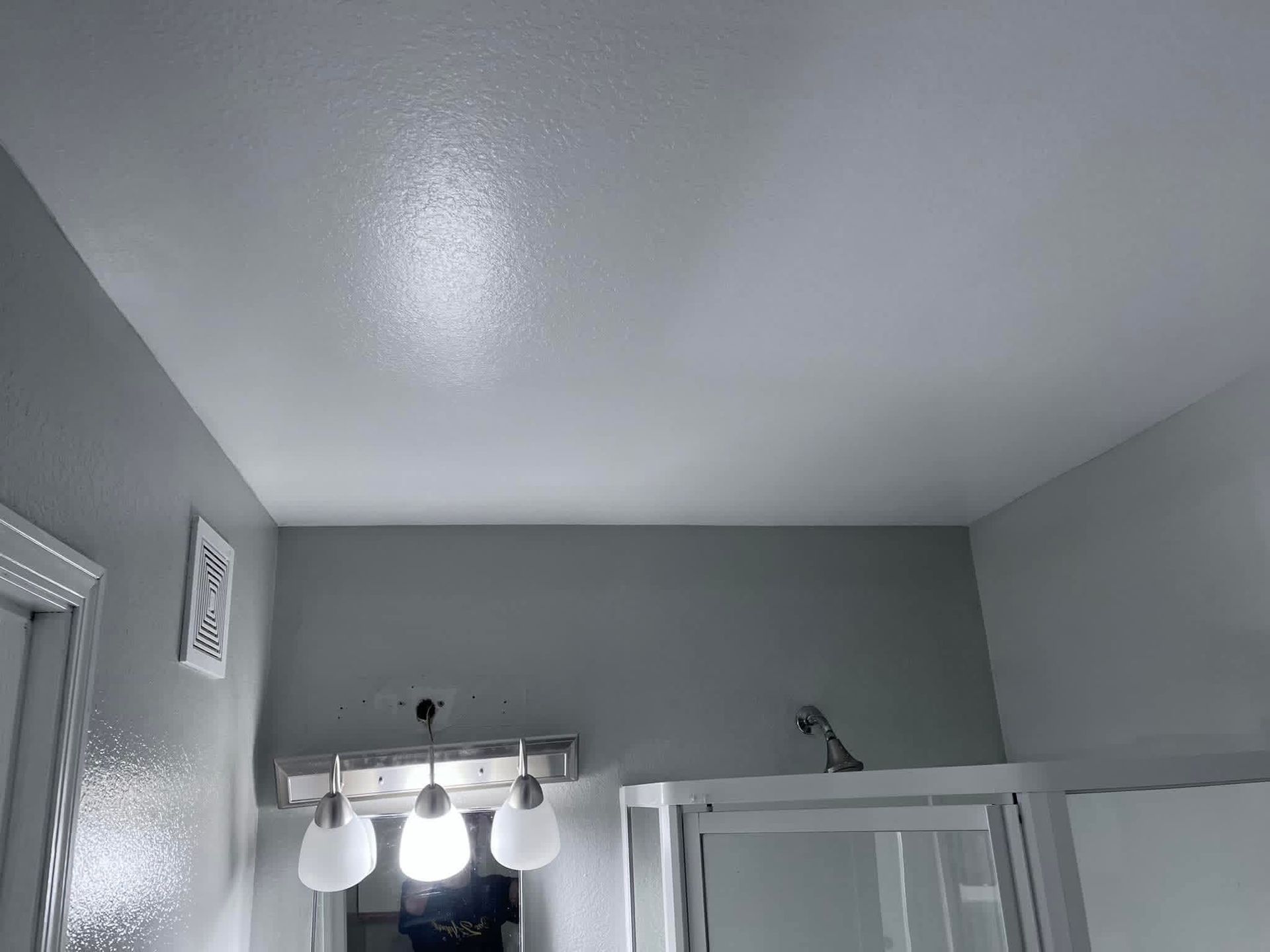 A bathroom interior featuring a light fixture with three white glass shades above a shower enclosure and gray walls.
