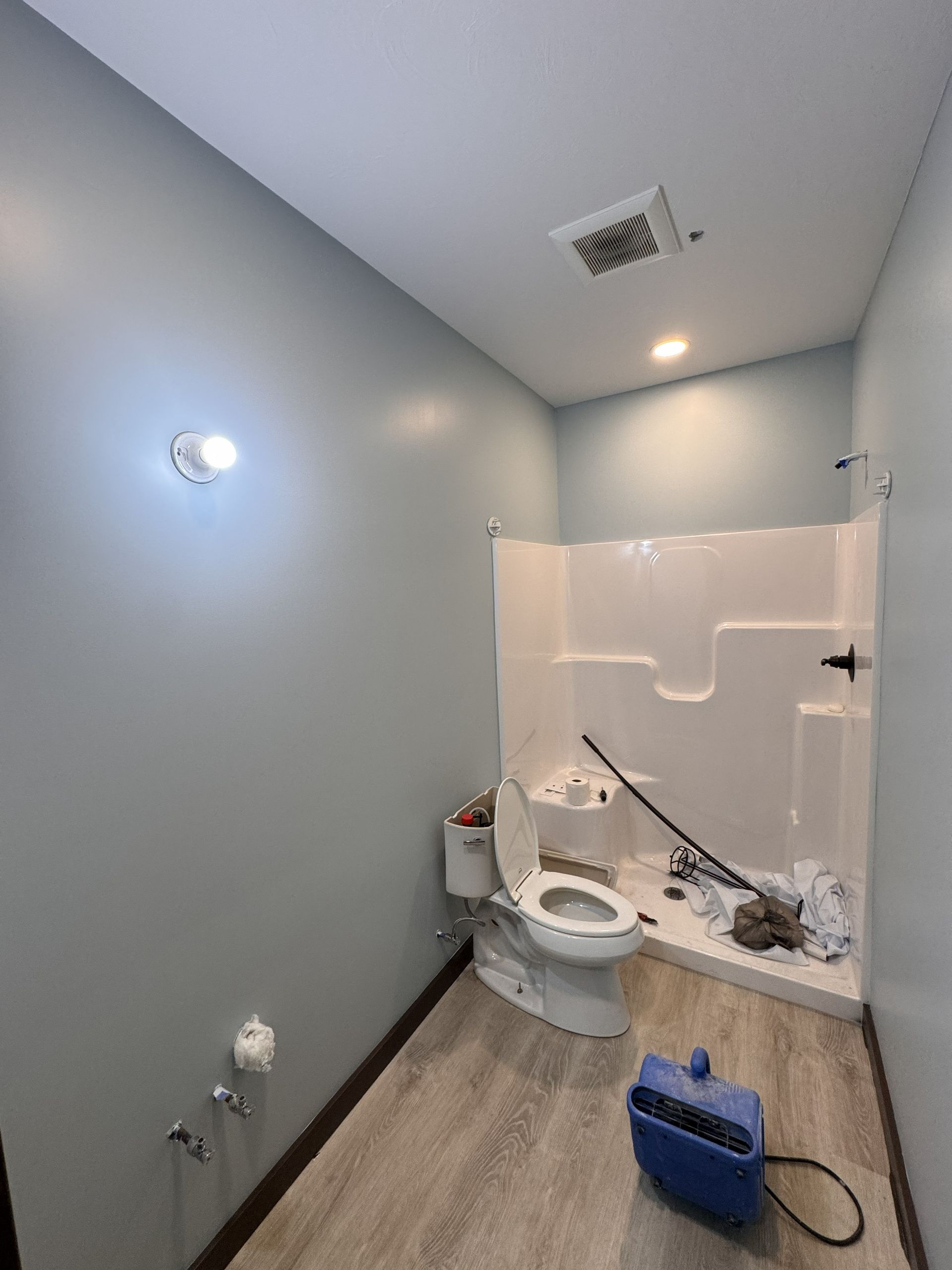 A light blue, unfinished bathroom with a white toilet and shower stall, light wood floors, and cleaning equipment.