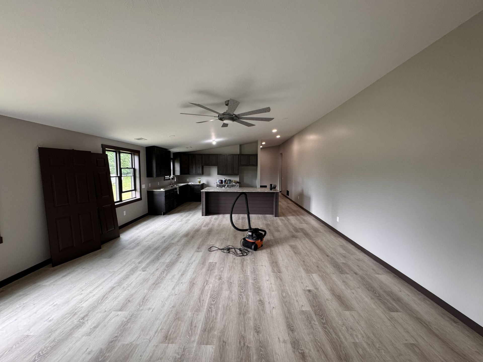 An open-concept living area with light wood floors, a dark kitchen island, and a vacuum cleaner in the center.