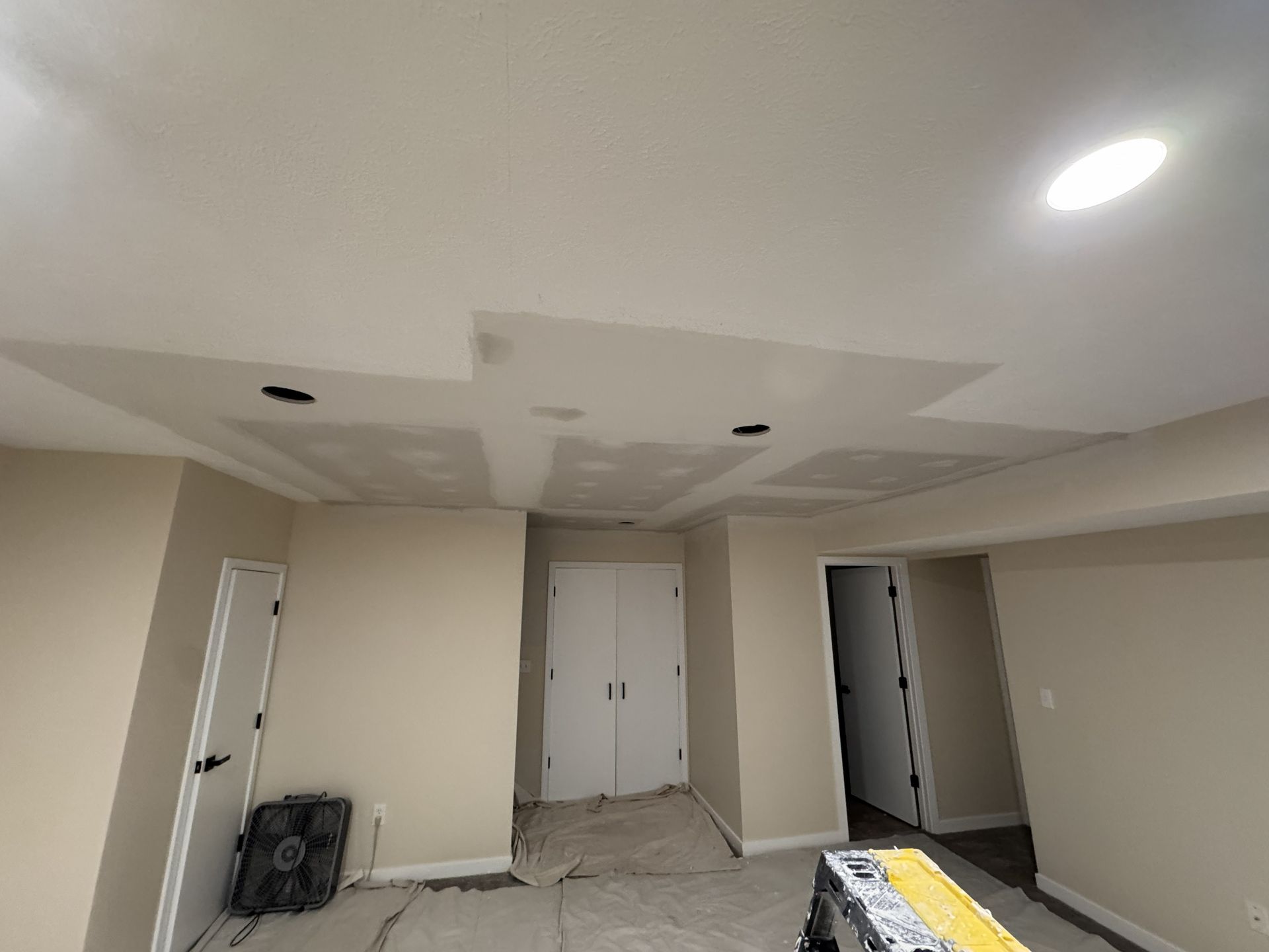 A beige room under renovation showing an unfinished drywall ceiling with patches, holes for light fixtures, and drop cloths.