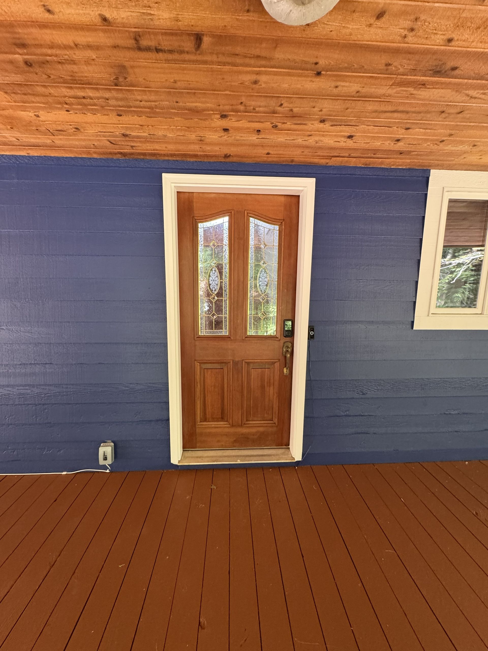 A brown wooden door with glass panels set into a vibrant blue exterior wall on a porch with brown decking.