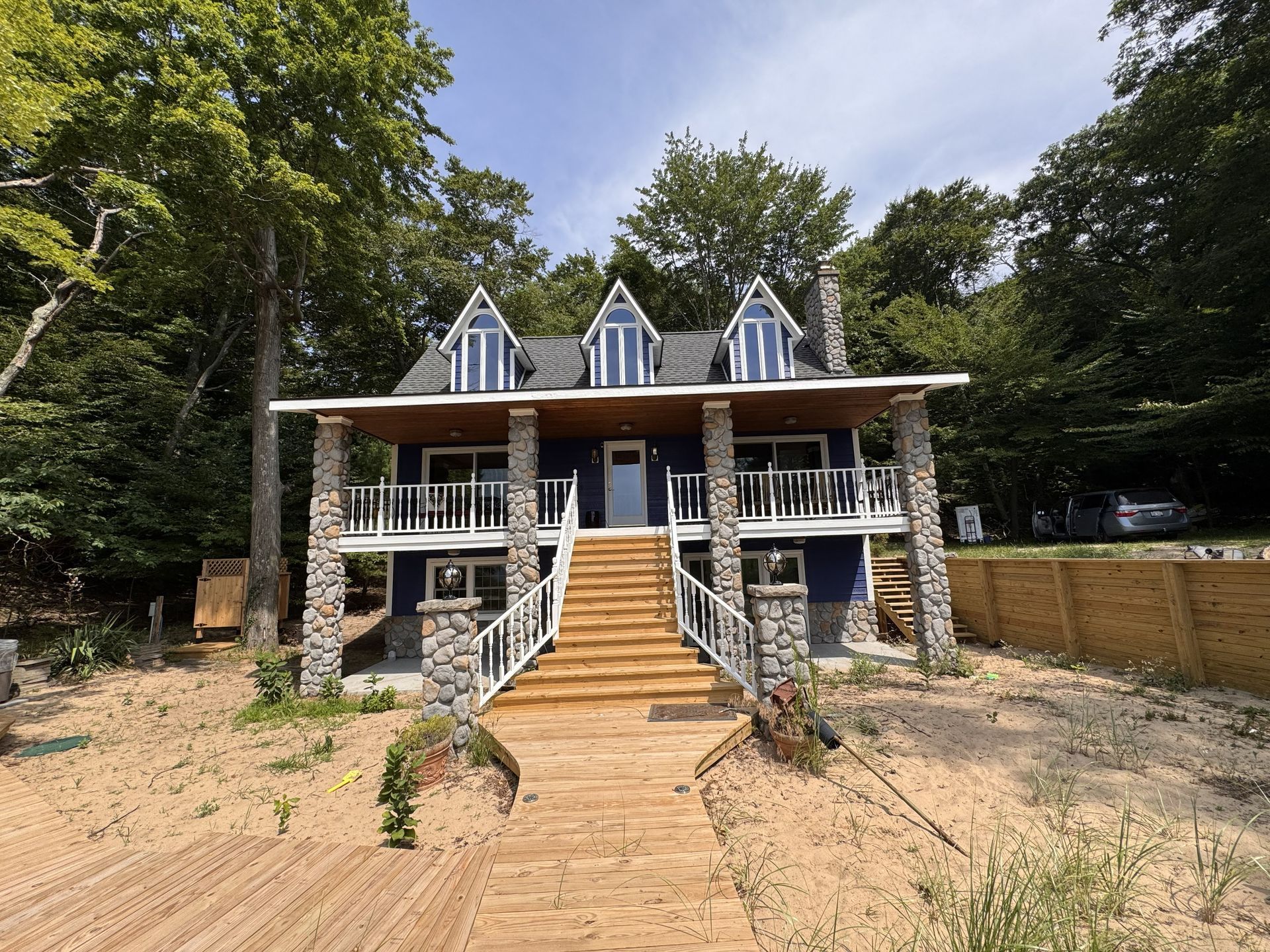 A blue, two-story house with a wooden staircase leading to the front porch, surrounded by sand and lush green trees.