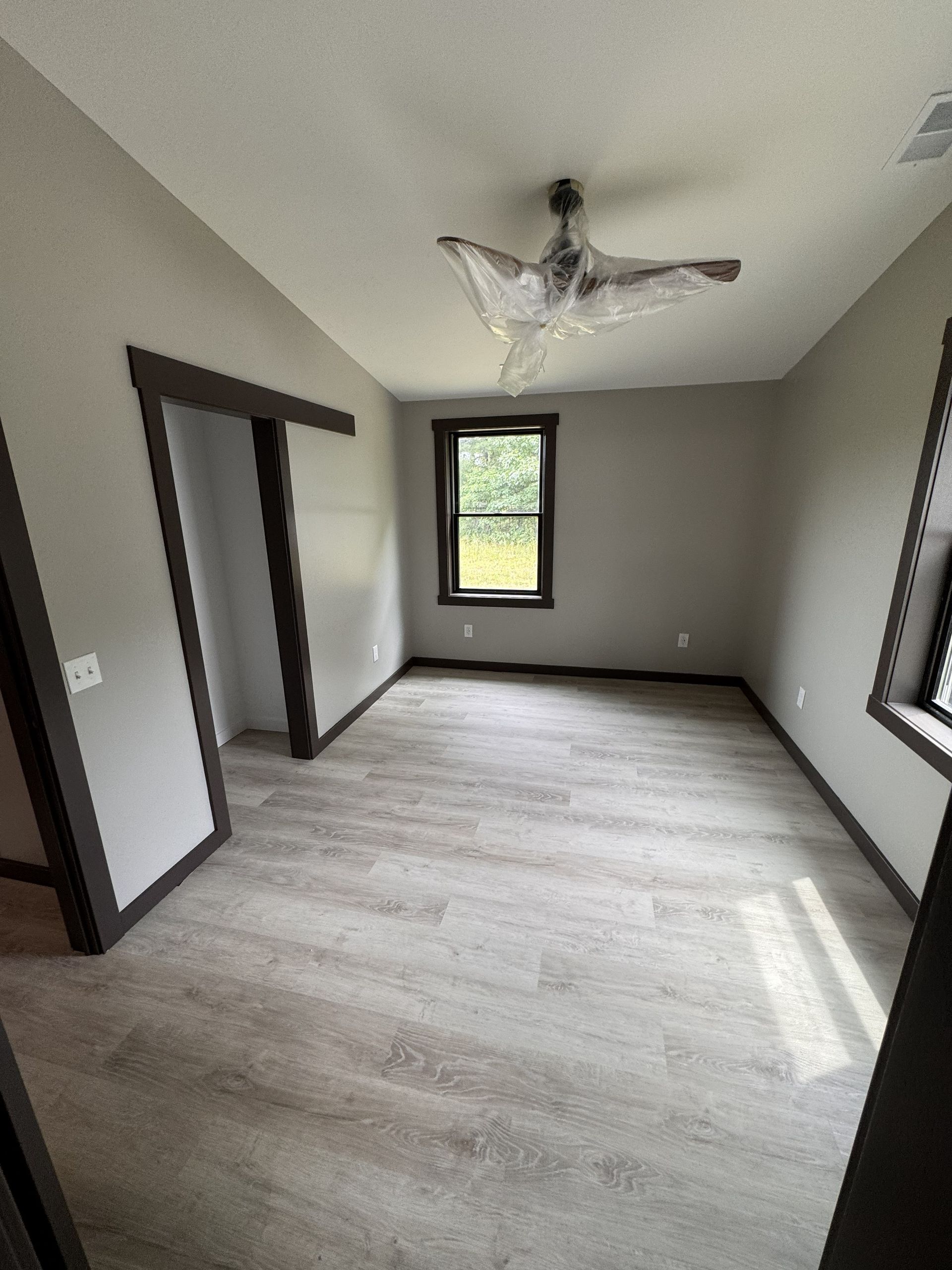 An empty room with light gray flooring, neutral-toned walls, dark trim, a single window, and a modern ceiling fan.
