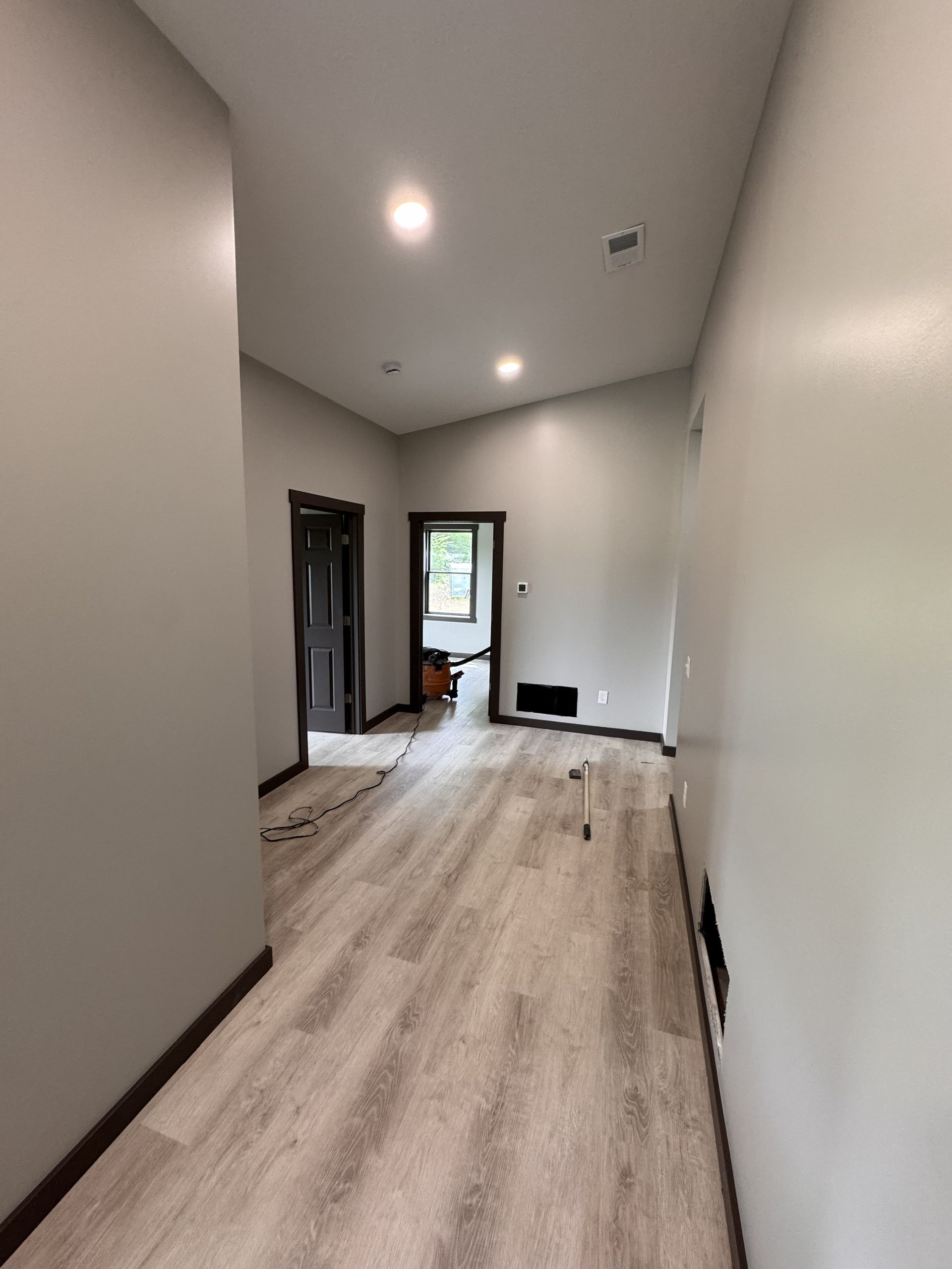 A hallway with wood-look flooring and light gray walls leads to a doorway showing a room with a window.