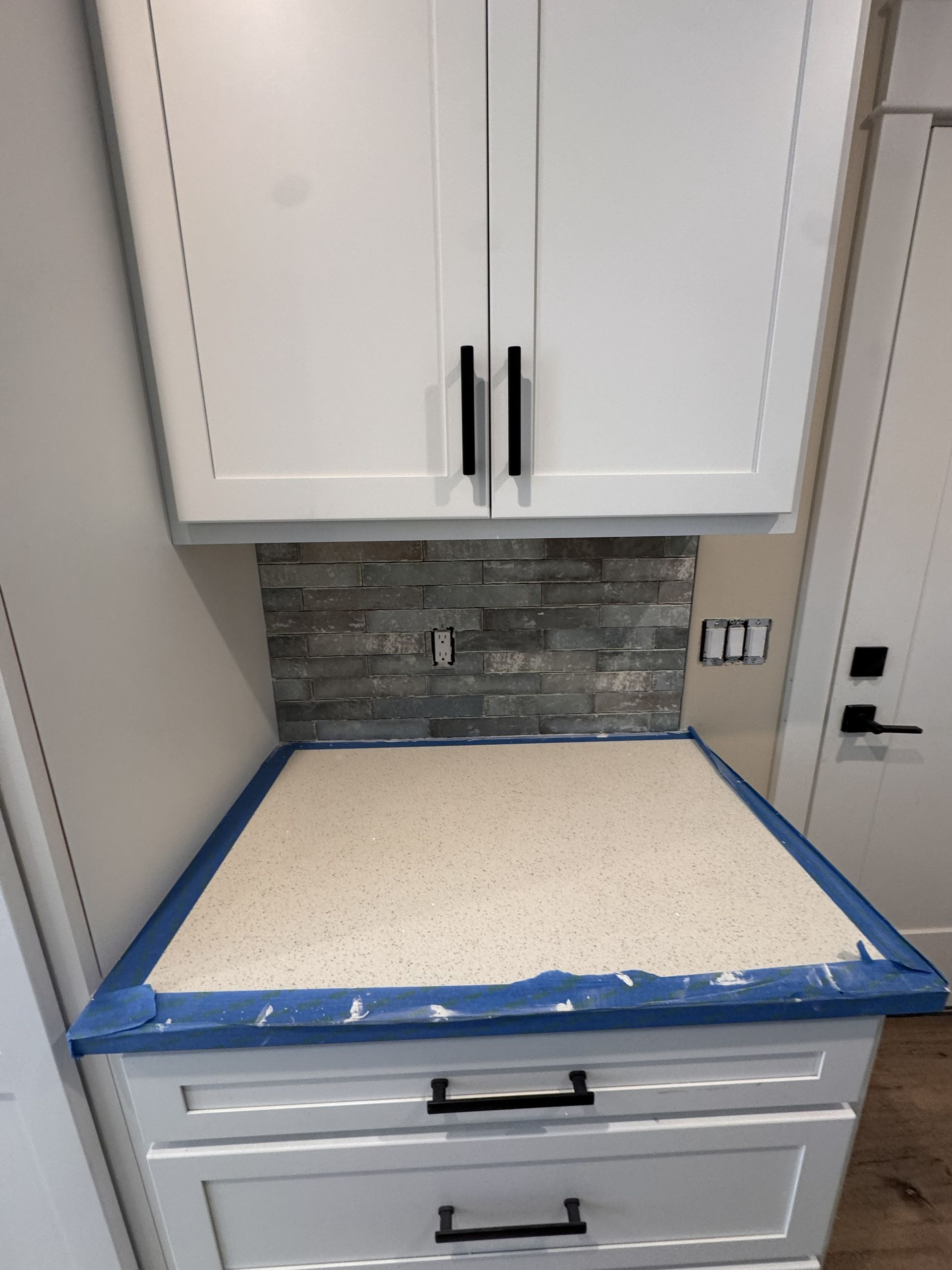 White cabinets with black handles, a stone backsplash, and a countertop covered with blue painter's tape in a kitchen.