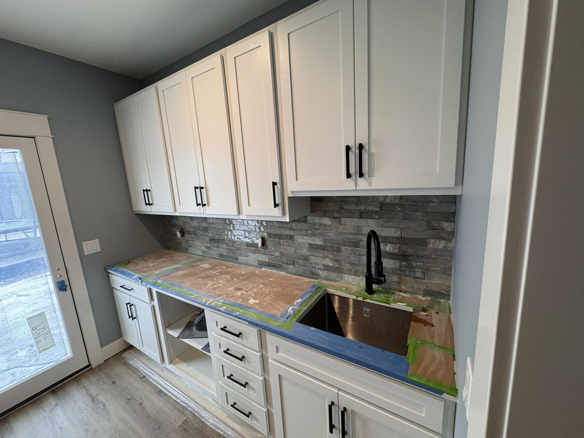 White kitchen cabinets and a stone backsplash with a sink installed in a room with light grey walls and a glass door.