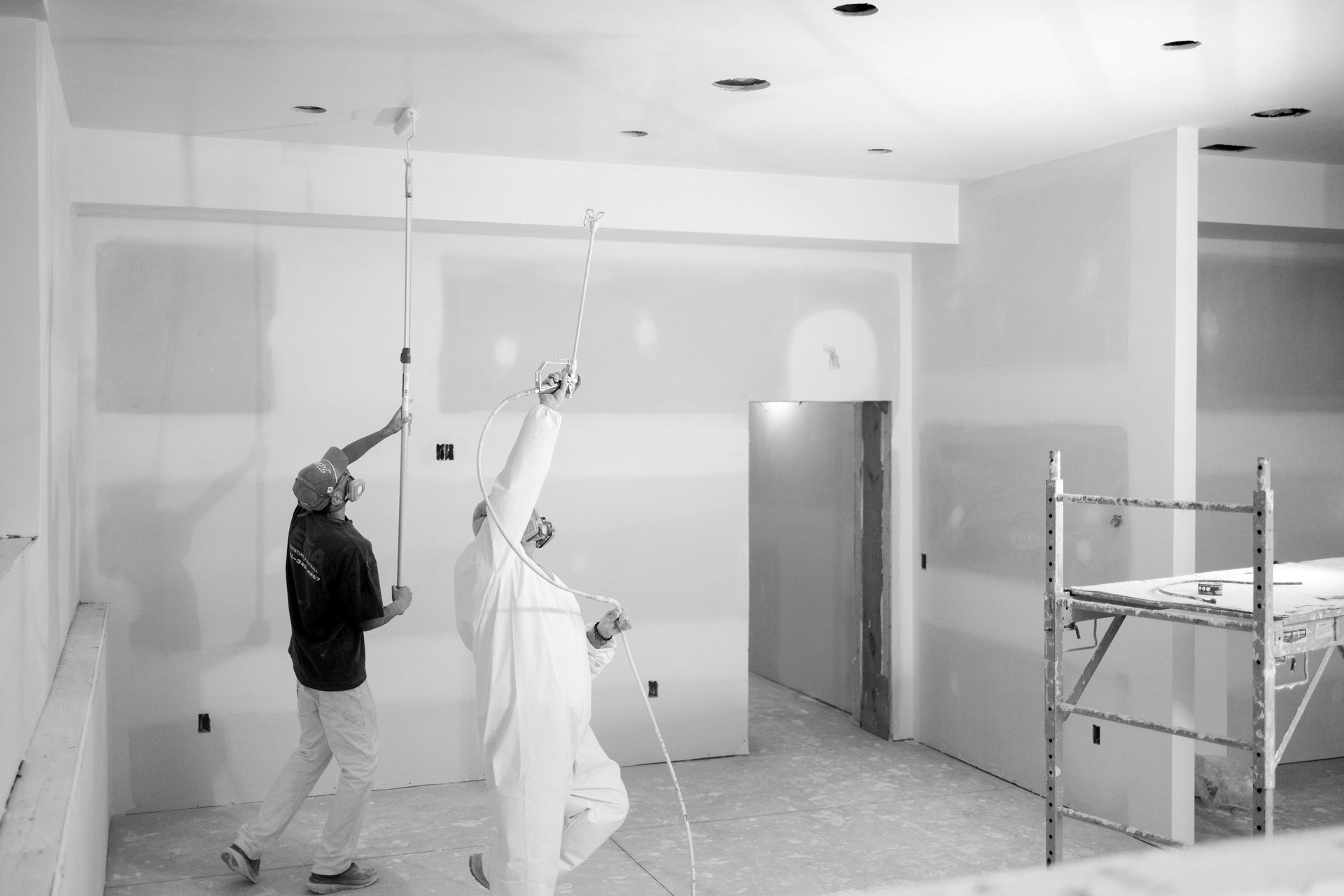 Two construction workers use paint rollers to prime the ceiling and walls of an unfinished room with white paint.