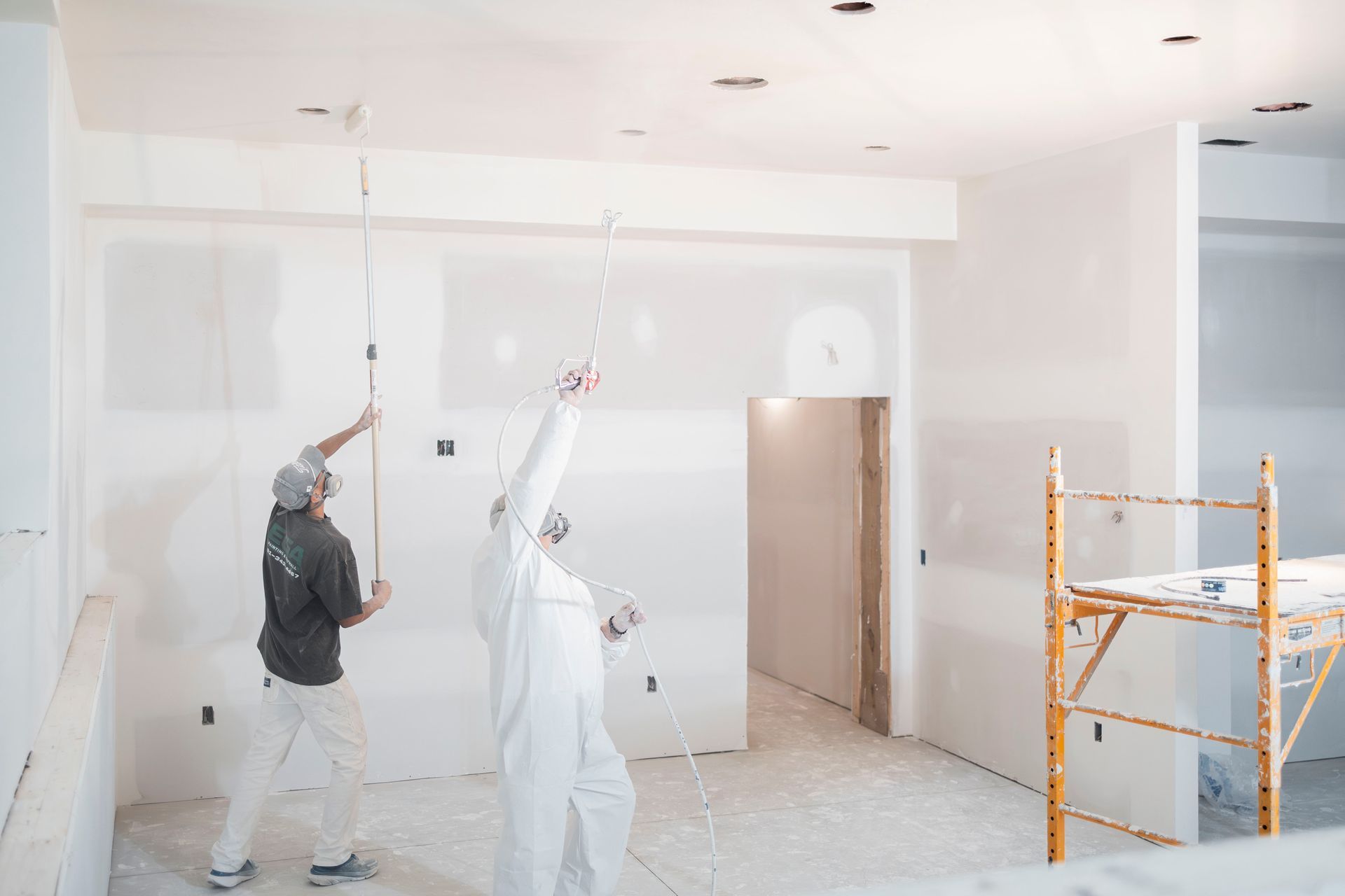 Two painters in protective gear work on the ceiling and walls of an unfinished room with a scaffold nearby.