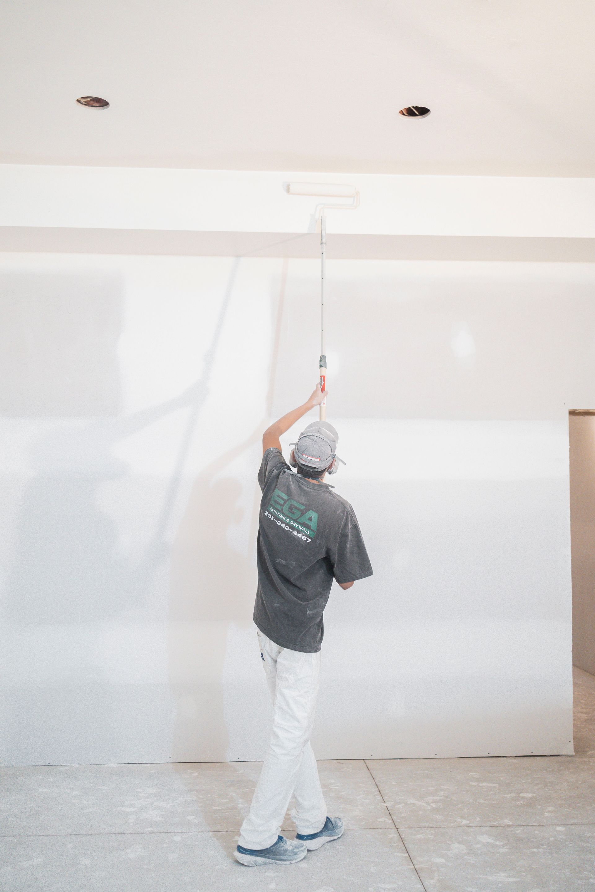 A person in a gray shirt and white pants uses a paint roller to paint a ceiling in an unfinished room.