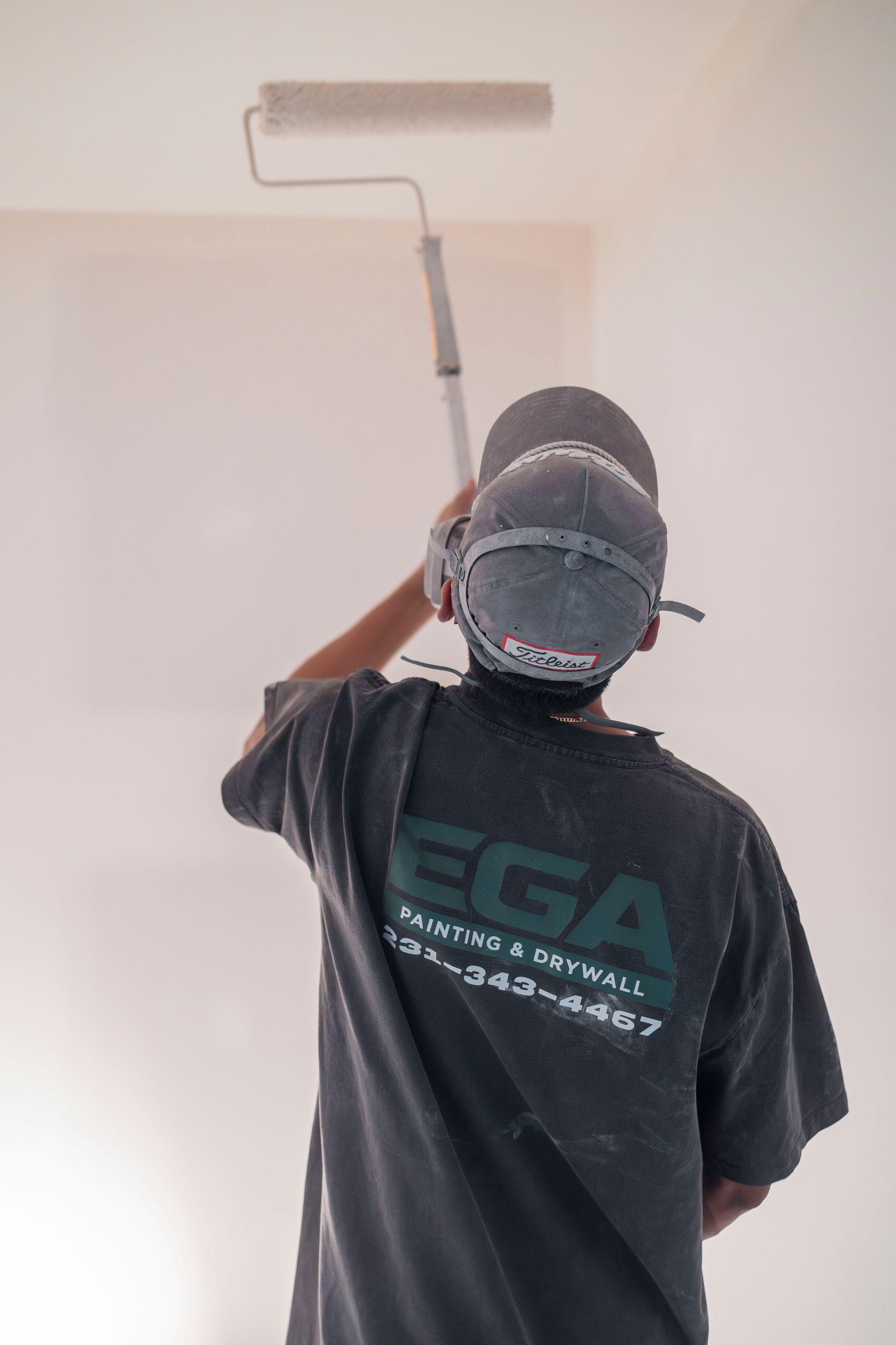 A person wearing a grey shirt and cap uses a roller on an extension pole to paint a white ceiling.